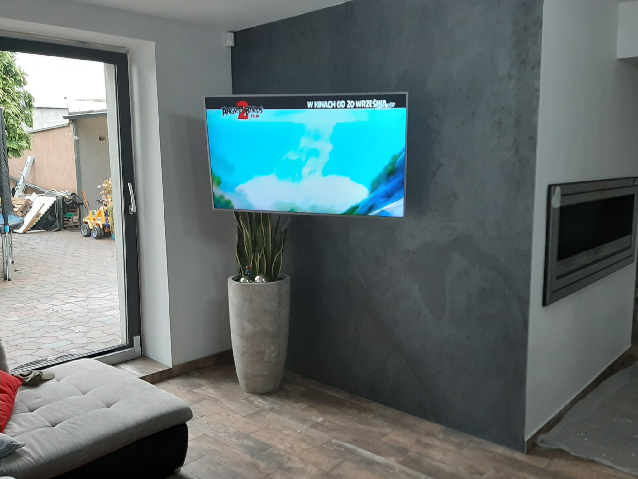 Salon z telewizorem wiszącym na ścianie z dekoracyjną szarą strukturą, obok kominek, pod telewizorem donica z rośliną, w tle widoczne okno.