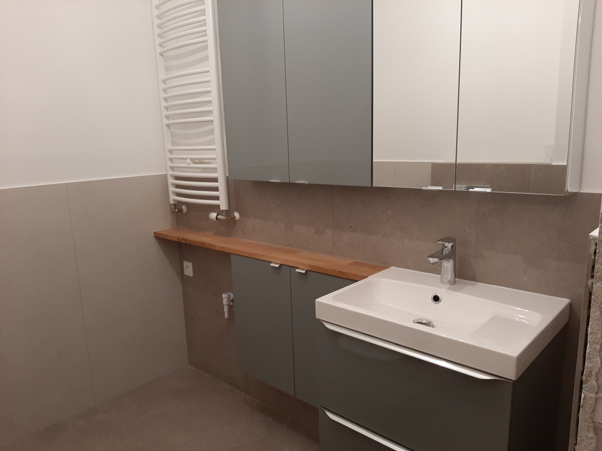 Nowoczesna łazienka po remoncie: szara zabudowa szafkowa pod umywalką z drewnianym blatem, biała umywalka, lustro w szafce wiszącej i grzejnik drabinkowy.