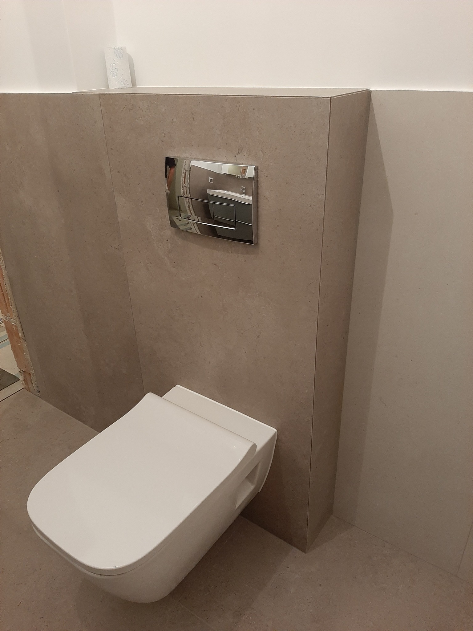 Nowoczesna toaleta podwieszana z chromowanym przyciskiem spłukującym, obudowana płytami ceramicznymi w beżowym odcieniu, z widoczną rolką papieru toaletowego na górnej krawędzi obudowy.