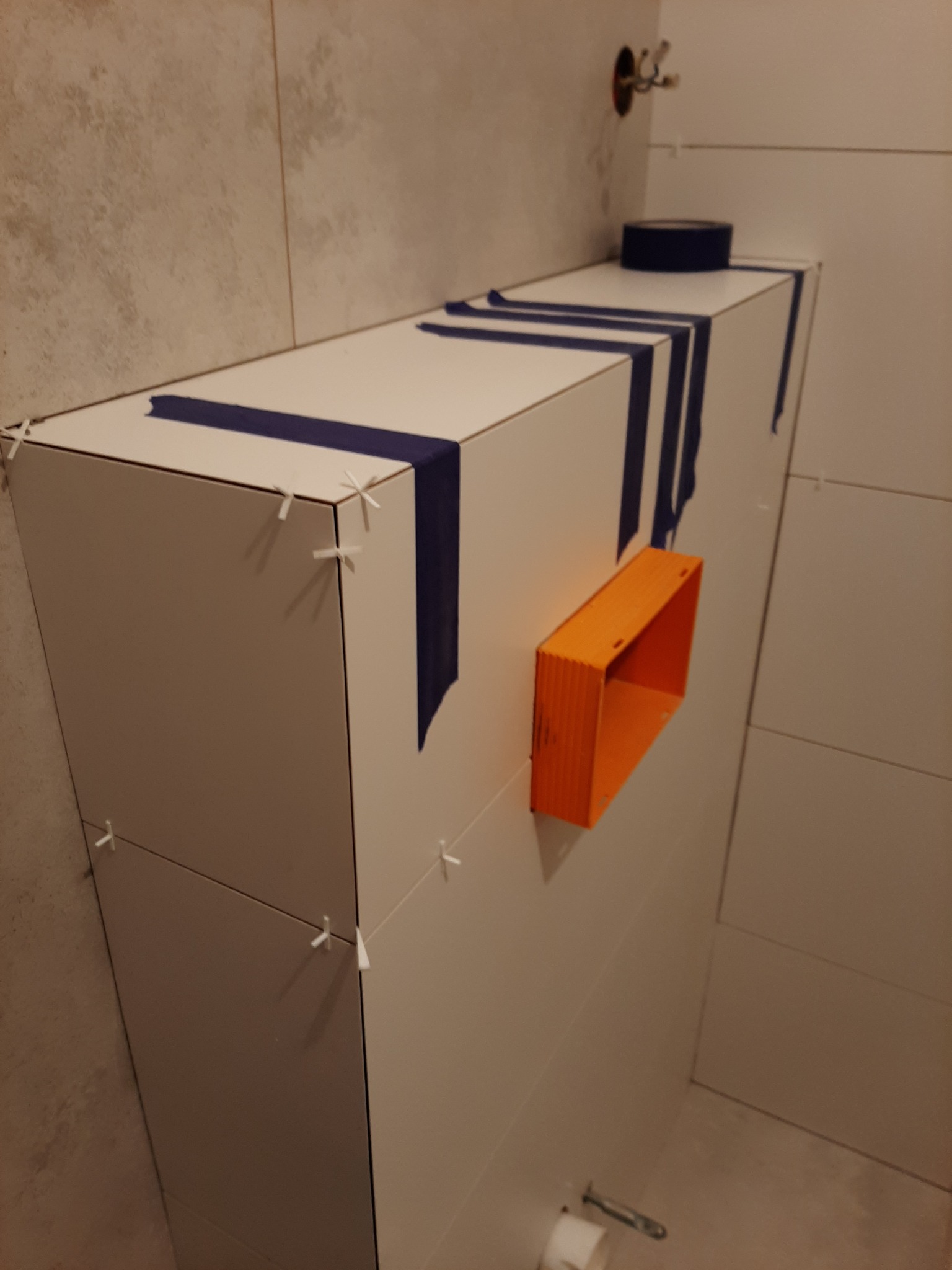 Biała zabudowa WC w trakcie wykańczania, z niebieską taśmą malarską, pomarańczową puszką instalacyjną i krzyżykami dystansowymi między płytkami.