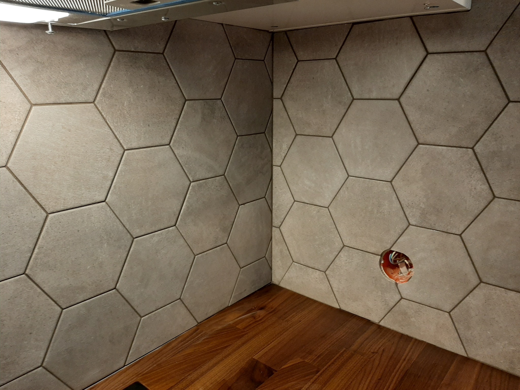 Fragment kuchni z heksagonalnymi płytkami ceramicznymi na ścianie i drewnianym blatem, widoczne gniazdko elektryczne w trakcie montażu.