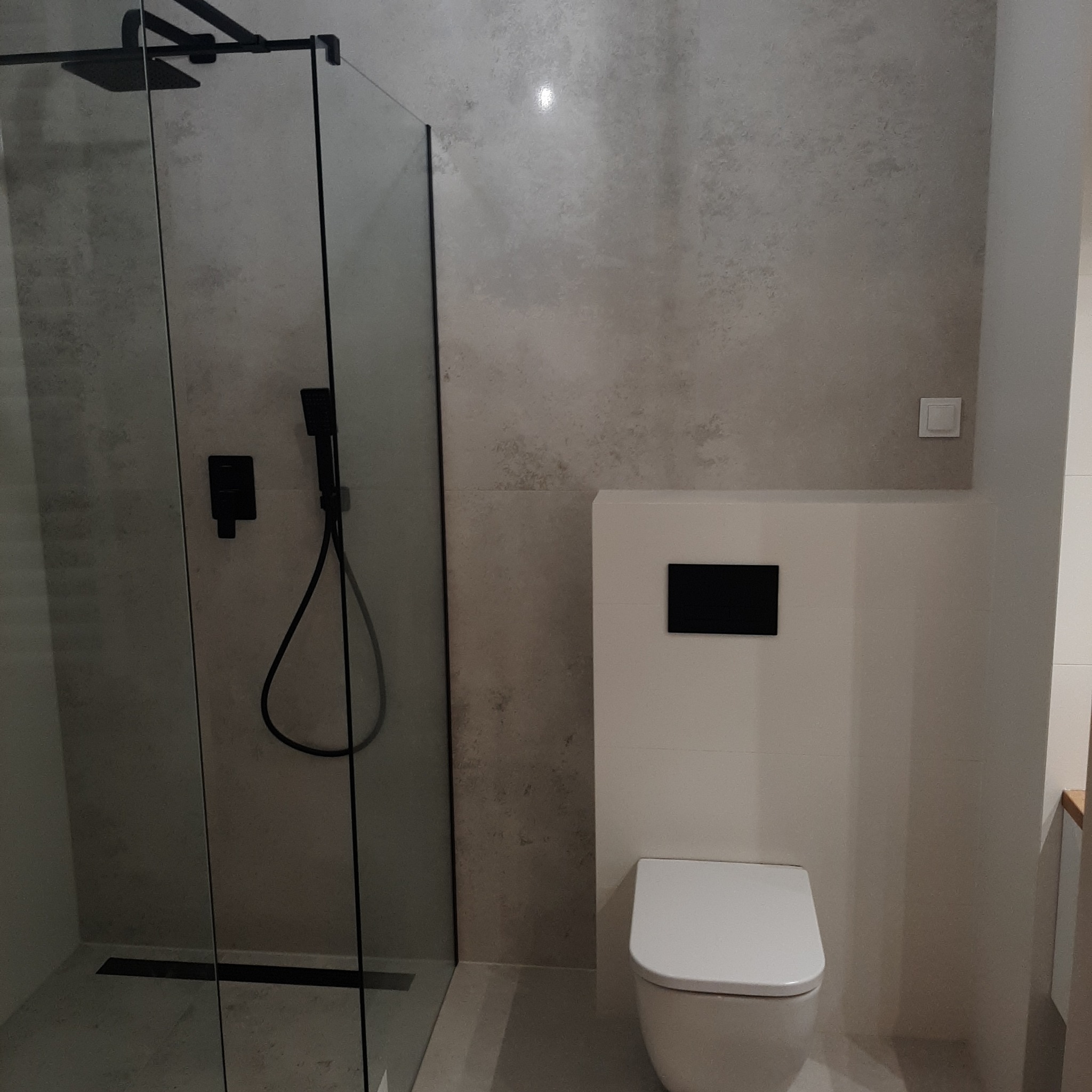 Nowoczesna łazienka z kabiną prysznicową typu walk-in z czarnymi elementami, szarą ścianą imitującą beton i podwieszaną toaletą z czarnym przyciskiem.
