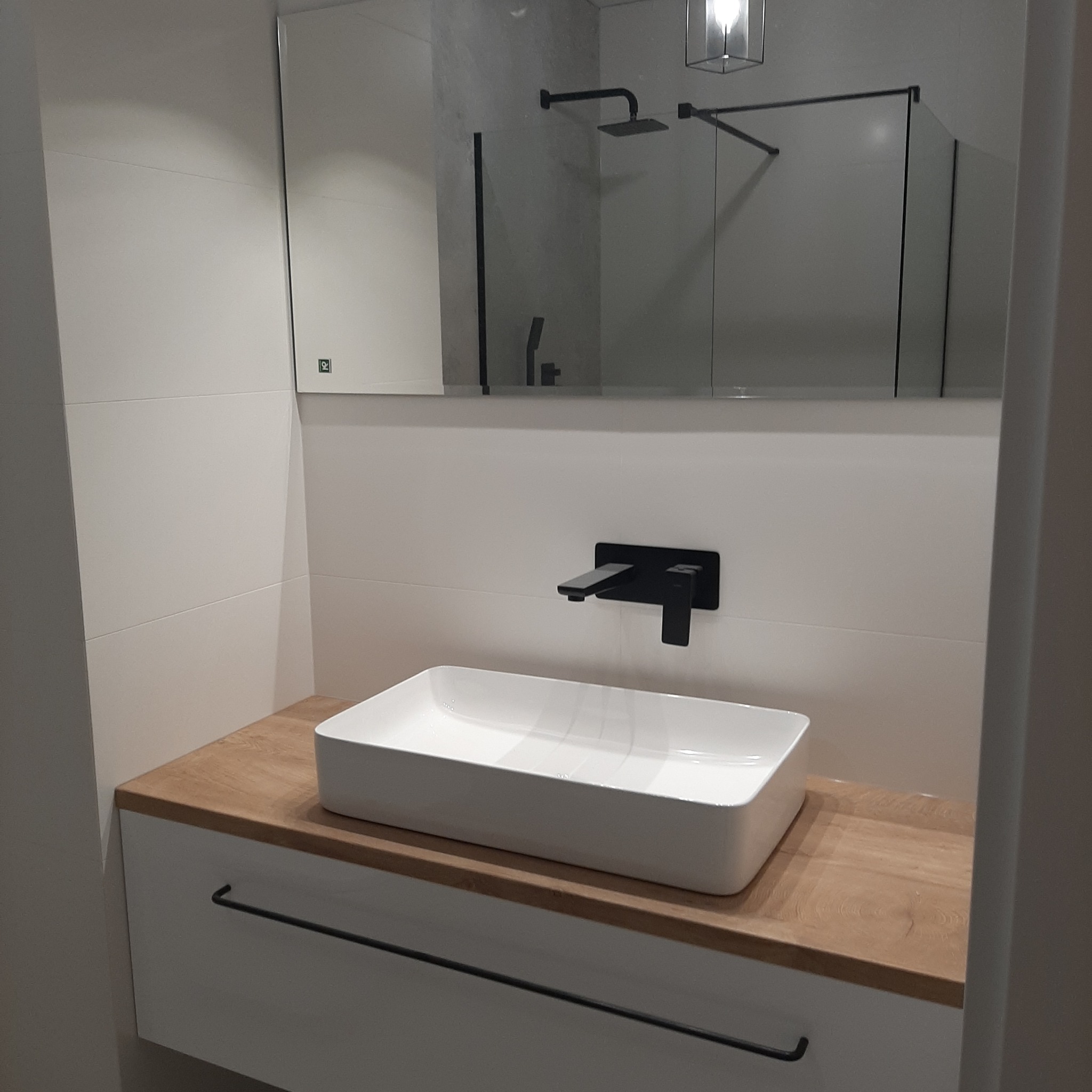 Nowoczesna łazienka z prostokątną białą umywalką nablatową, czarną armaturą i lustrem odbijającym kabinę prysznicową z czarnymi ramami.