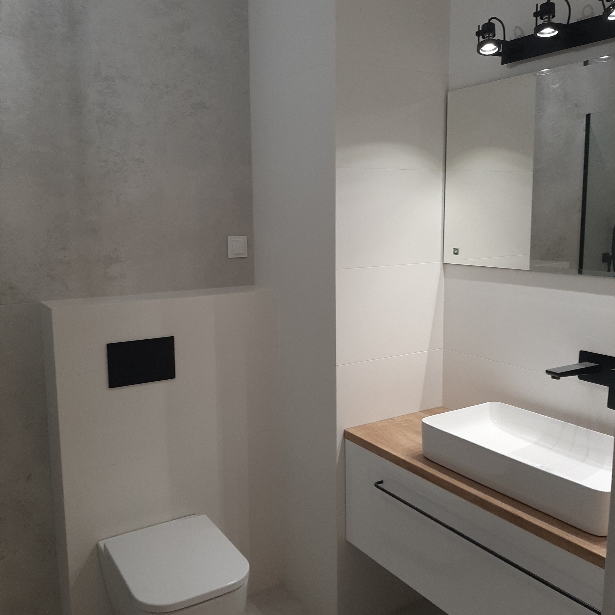 Nowoczesna łazienka z umywalką nablatową, czarną armaturą i szarą ścianą imitującą beton.