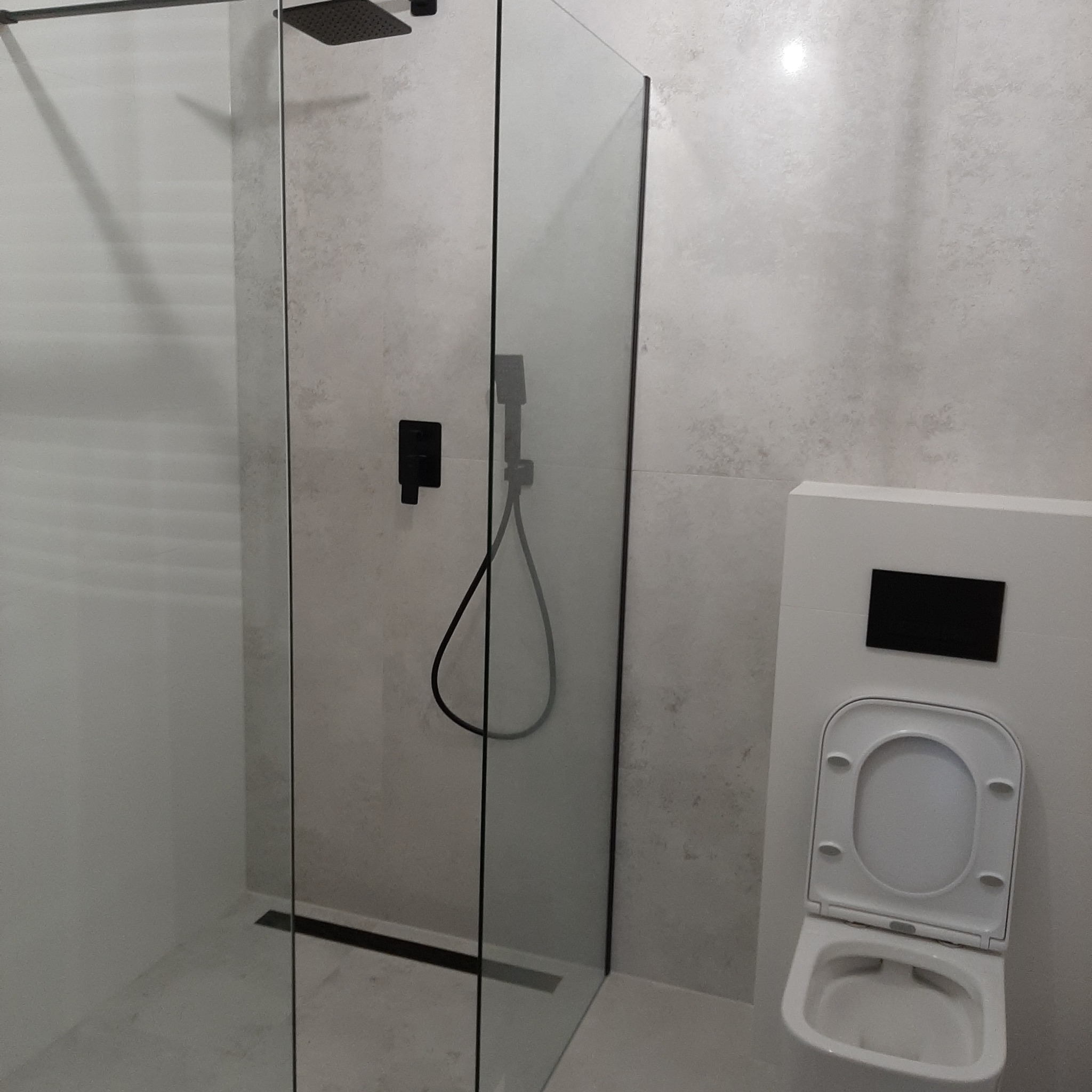 Nowoczesna kabina prysznicowa ze szklaną ścianką i czarnymi elementami armatury, widok na podwieszaną toaletę z białą miską i czarnym przyciskiem spłukiwania, jasne płytki na ścianach.