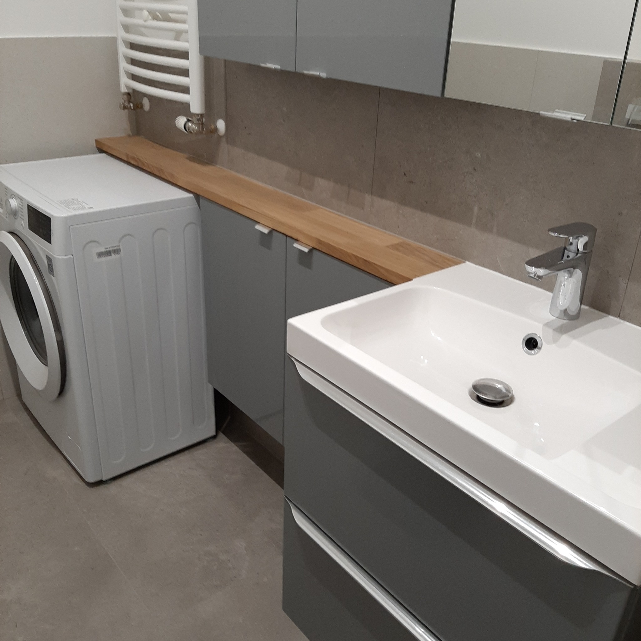 Nowoczesna łazienka z szarą zabudową, drewnianym blatem, białą umywalką i pralką, minimalistyczny design.