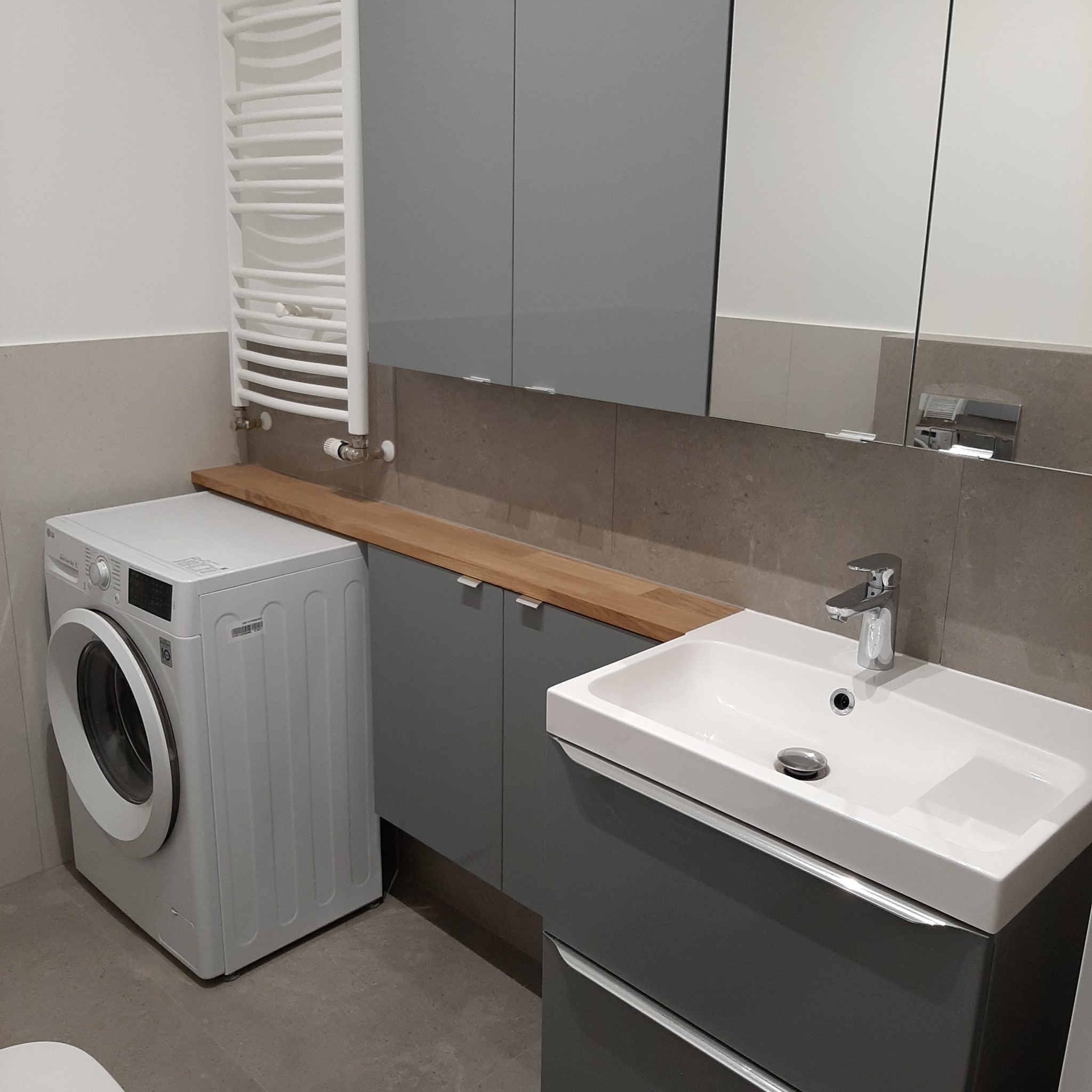 Nowoczesna łazienka w szarościach z pralką, umywalką z szafką i grzejnikiem drabinkowym. Drewniany blat nad szafkami dodaje ciepła wnętrzu.