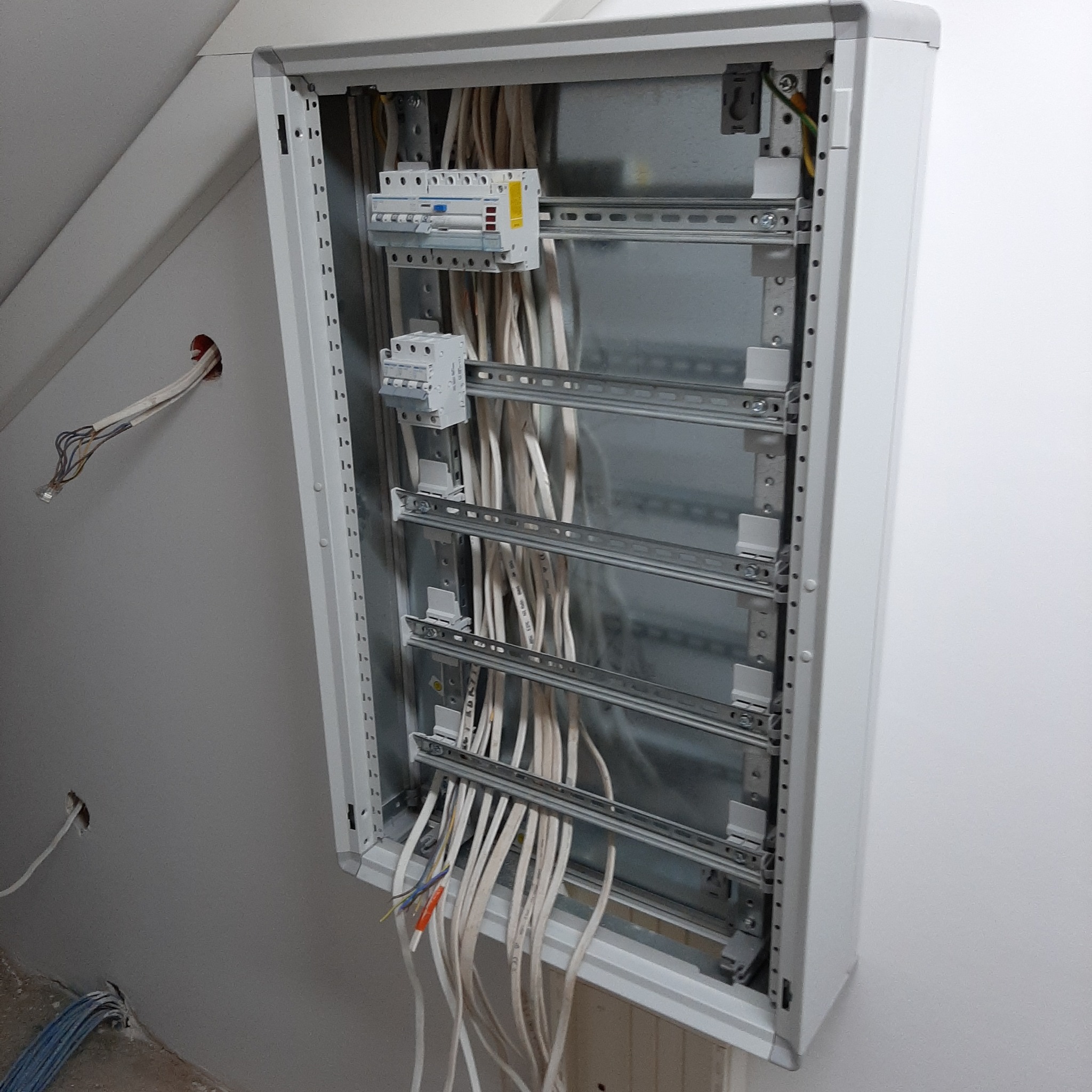 Otwarta, biała skrzynka elektryczna zamontowana na ścianie pod skosem, z widocznymi zabezpieczeniami i licznymi przewodami elektrycznymi wewnątrz oraz wystającymi z dołu i góry, część przewodów...