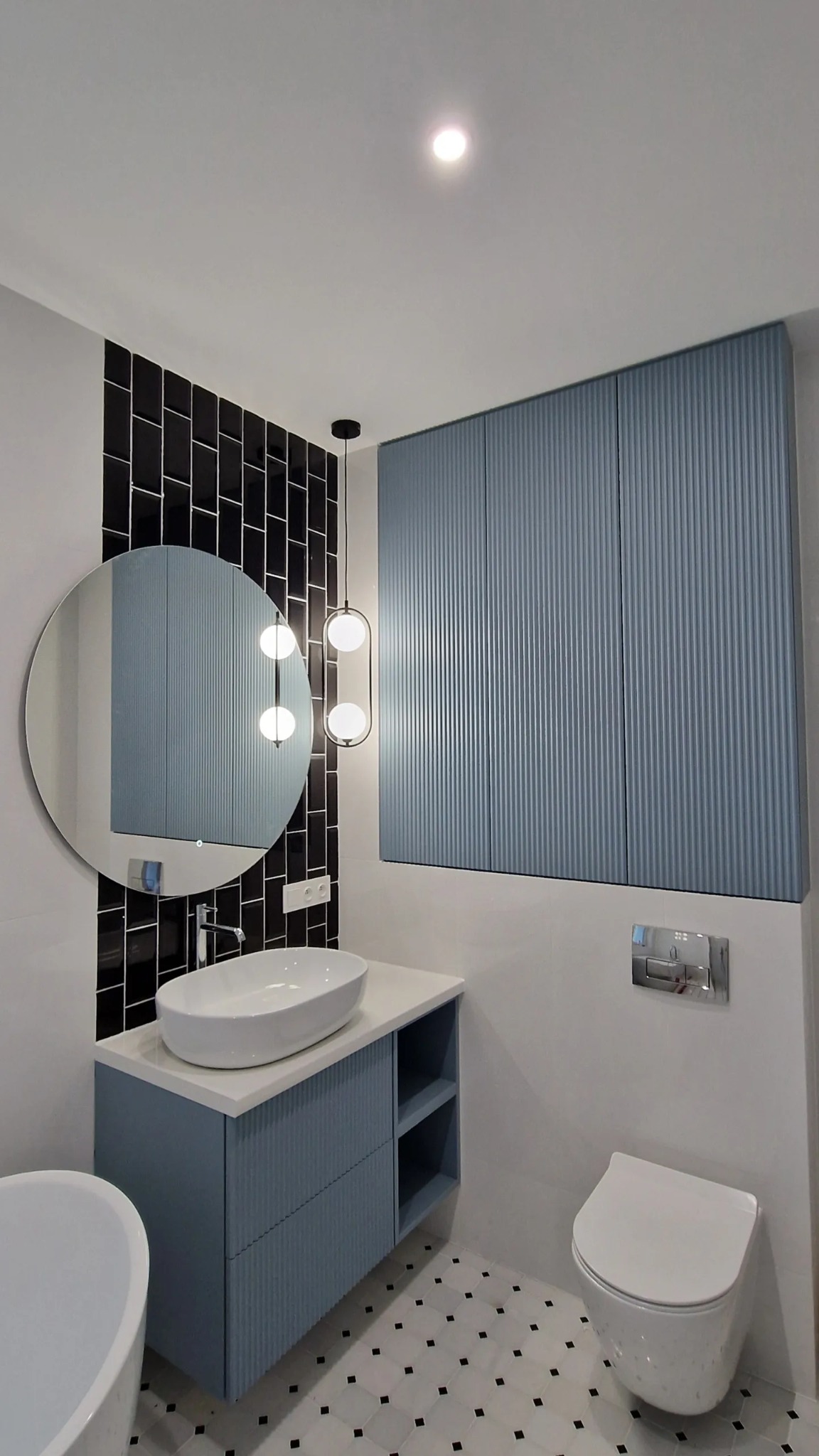 Niebieska szafka łazienkowa z umywalką nablatową, wisząca szafka z ryflowanymi frontami, okrągłe lustro z podświetleniem LED i czarne płytki typu metro ułożone pionowo w nowoczesnej łazience.