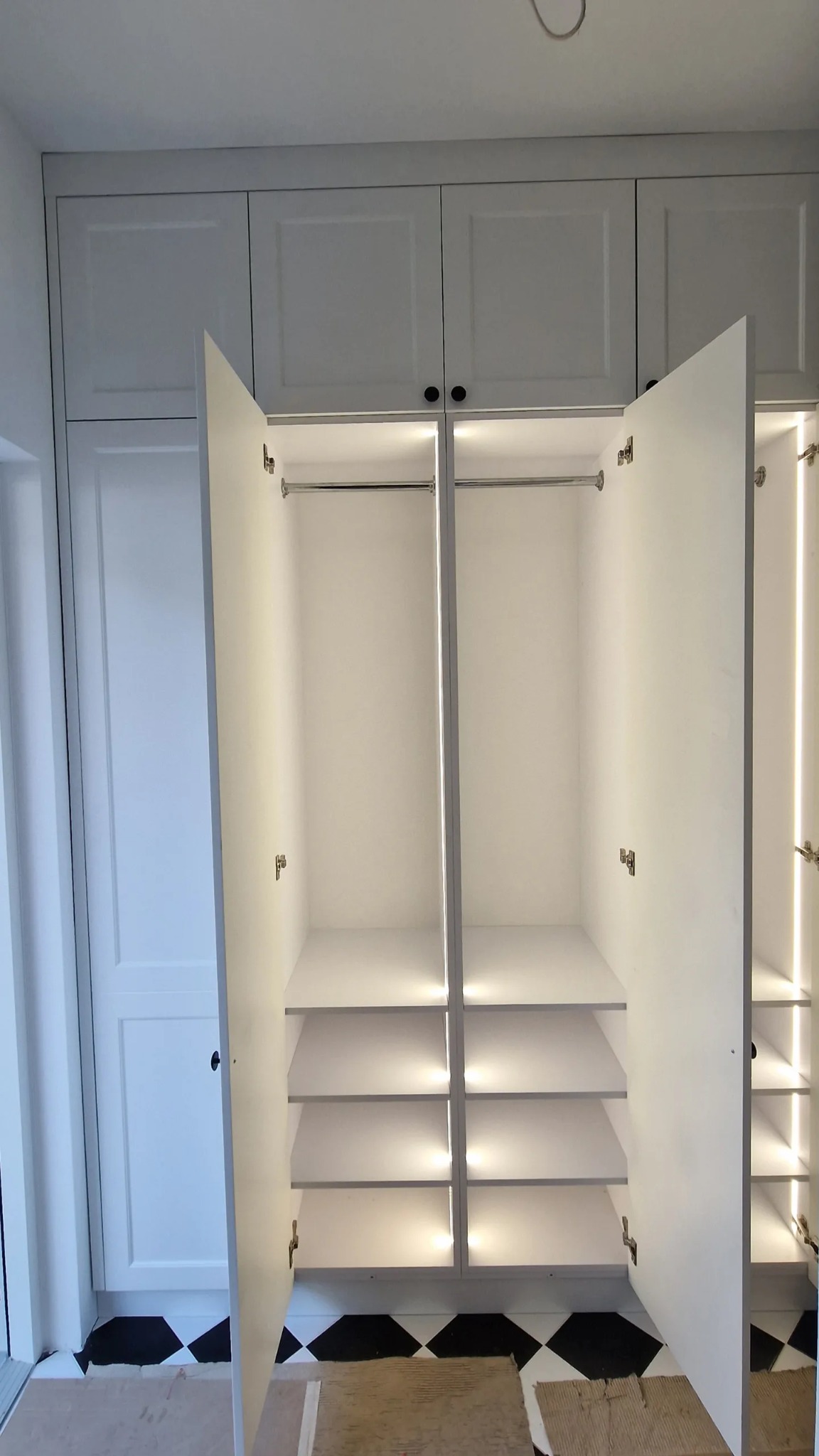 Biała szafa wnękowa z otwartymi drzwiami, oświetleniem LED, półkami i drążkami, widoczna na tle czarno-białej podłogi w romby.