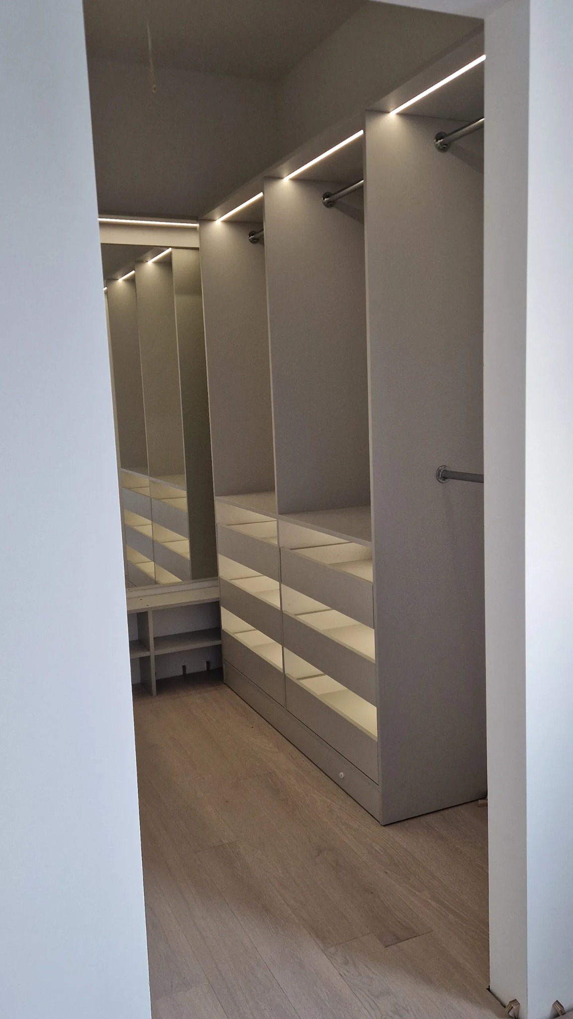 Wnętrze garderoby z oświetleniem LED w półkach i nad drążkami, widok zza rogu ściany, drewniana podłoga.
