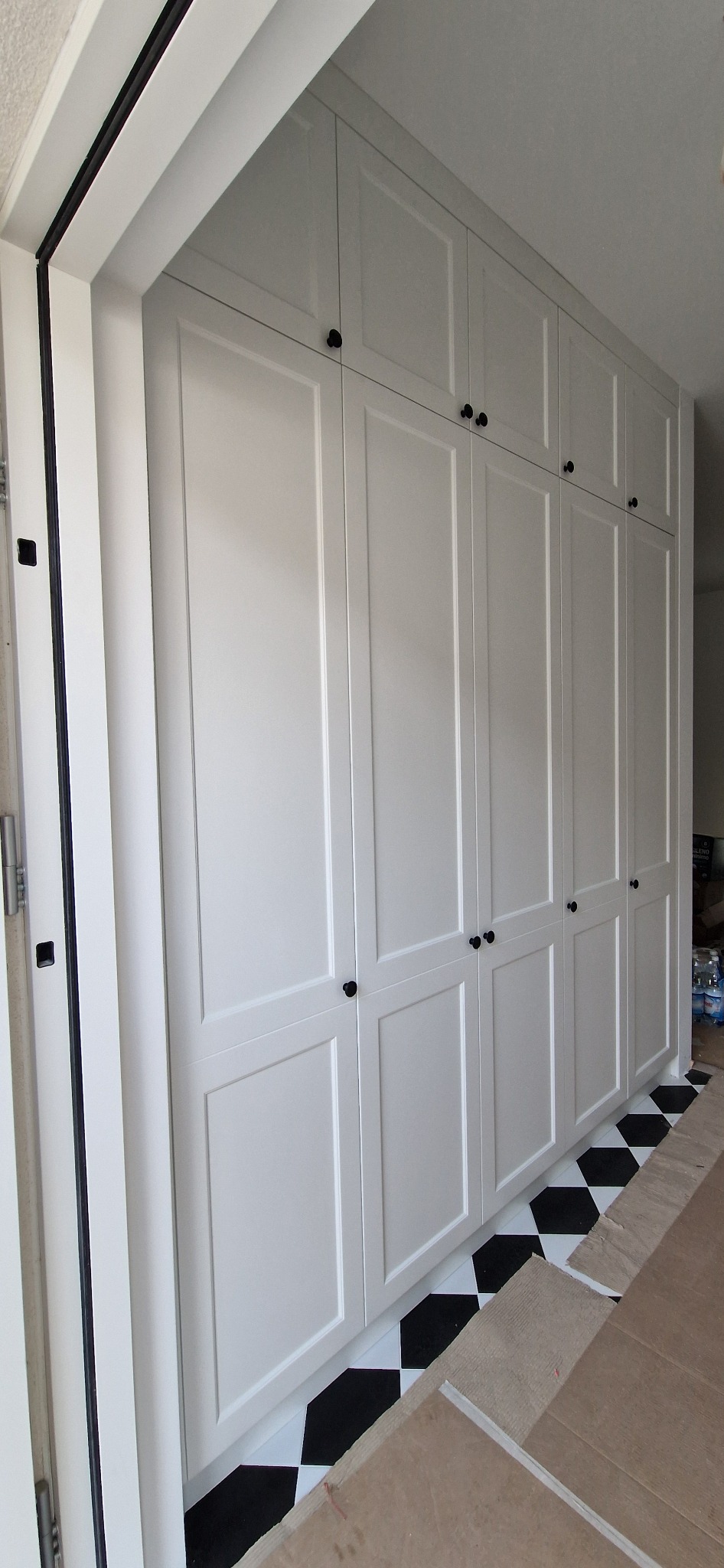 Wysoka, biała szafa wnękowa z czarnymi gałkami, widoczna z boku, z fragmentem czarno-białej podłogi w szachownicę i niezakończonym wykończeniem podłogi.