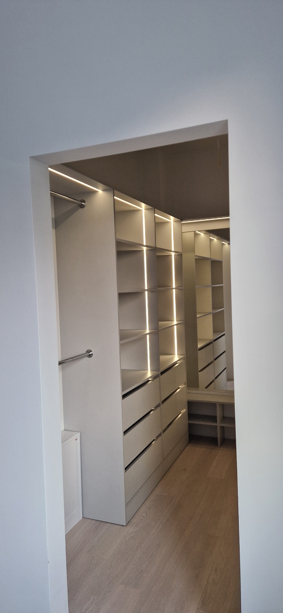 Nowoczesna, podświetlana garderoba z szufladami i półkami, widoczna z korytarza, z drewnianą podłogą.