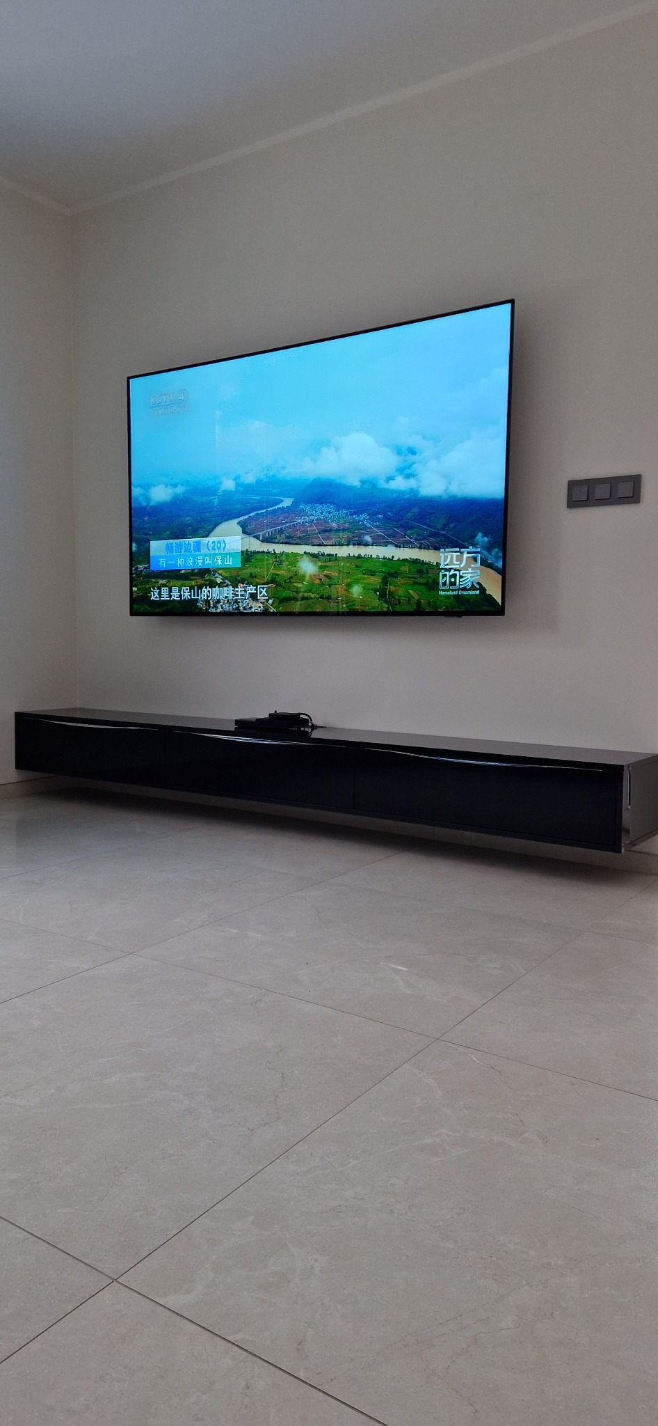 Długa, czarna szafka RTV podwieszona pod telewizorem na ścianie, minimalistyczny design, jasna podłoga z płytek.