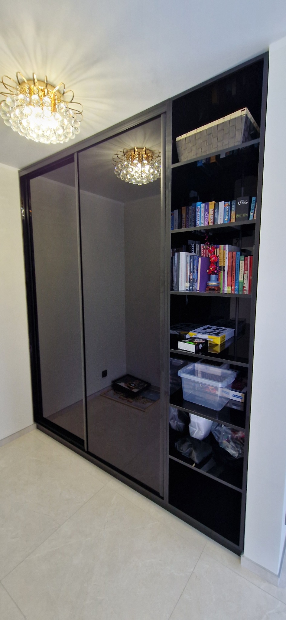 Czarna szafa wnękowa z przesuwnymi, lustrzanymi drzwiami i otwartymi półkami, wypełniona książkami i pudełkami, oświetlona dekoracyjnym plafonem.