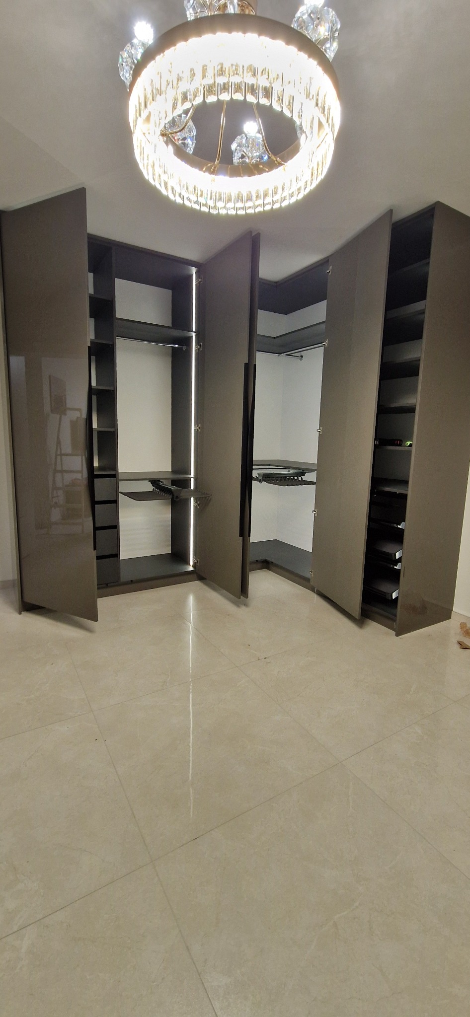 Przestronna garderoba narożna z szarymi frontami, oświetleniem LED wewnątrz i dekoracyjnym żyrandolem kryształowym.