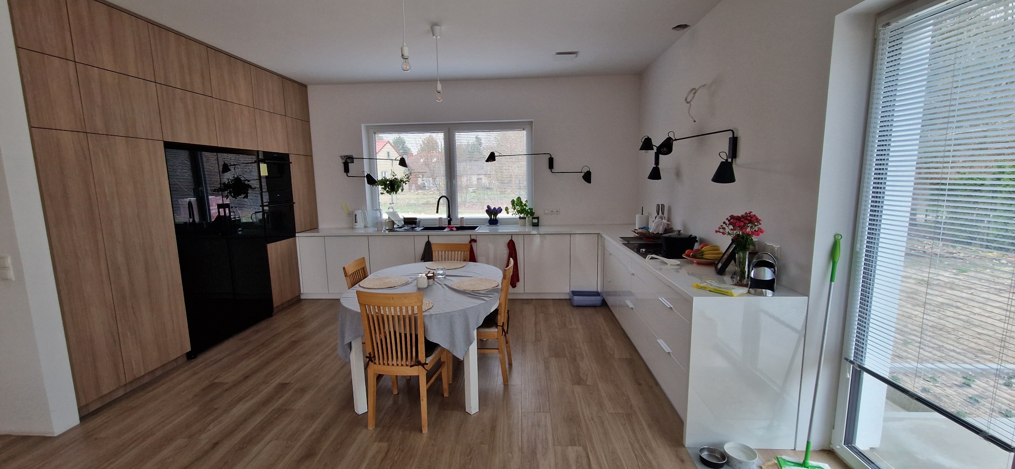 Jasna kuchnia z drewnianymi szafkami, czarnym sprzętem AGD, białymi blatami i owalnym stołem z szarym obrusem, oświetlona lampami w stylu industrialnym.