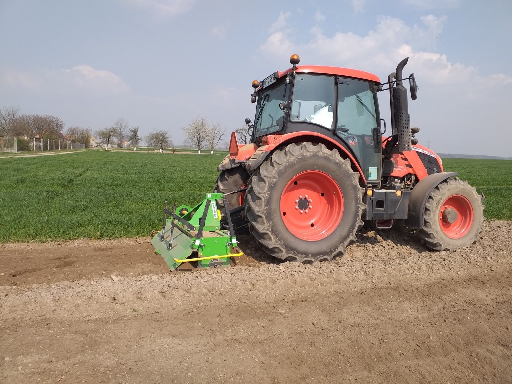 Czerwony traktor Kubota z zielonym agregatem uprawowym przygotowuje pas ziemi na polu uprawnym, widoczne ślady pracy i zielone zboże w tle.