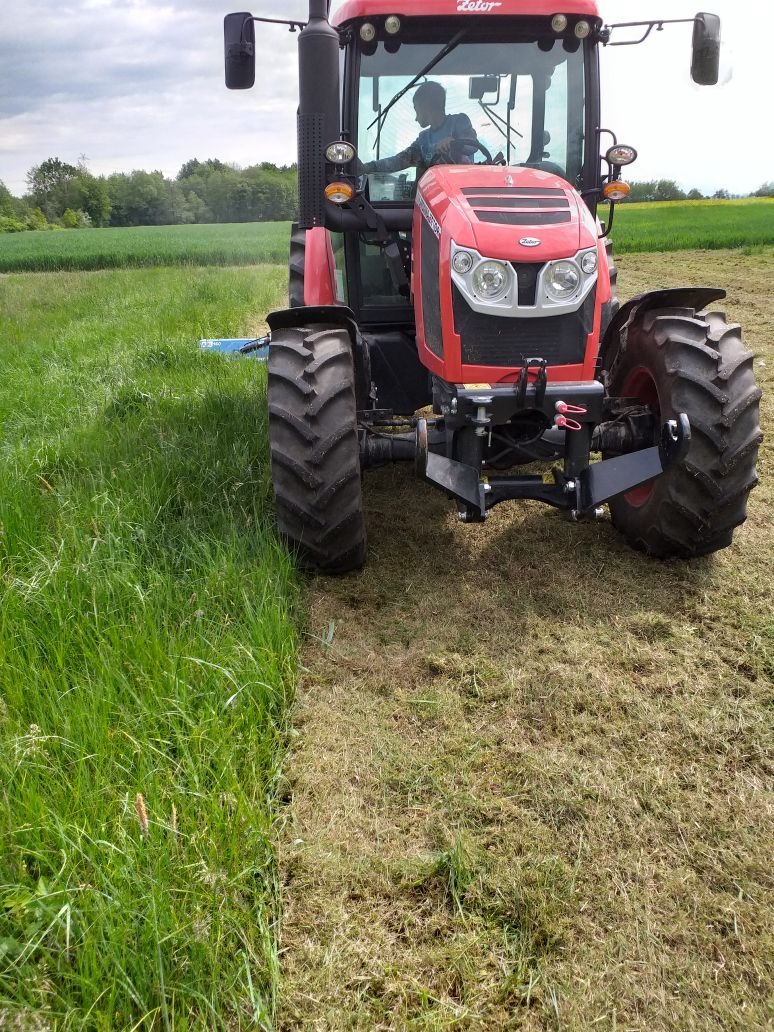 Czerwony traktor Zetor z operatorem podczas koszenia wysokiej trawy na polu, widoczna linia cięcia, tło stanowią drzewa i pochmurne niebo.