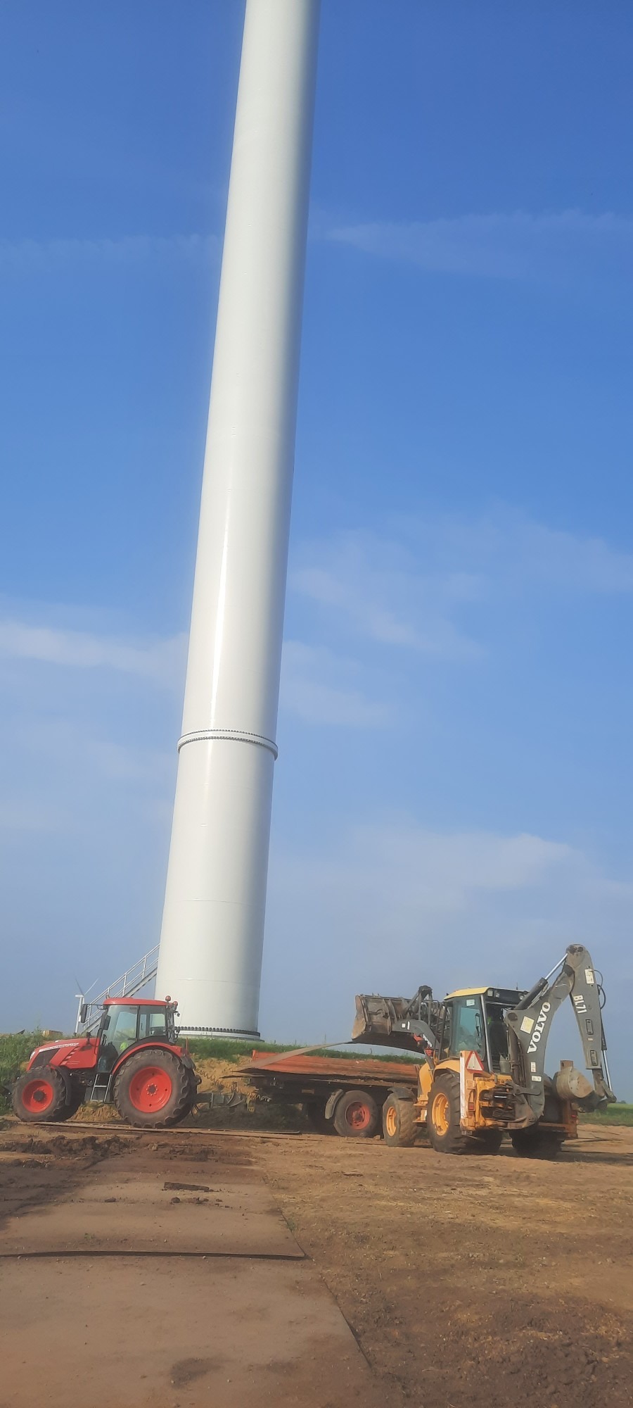 Czerwony traktor i żółta koparko-ładowarka Volvo BL71 pracują przy montażu segmentu białej turbiny wiatrowej na tle błękitnego nieba z delikatnymi chmurami.