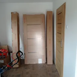 Przygotowane do montażu drewniane drzwi wewnętrzne w opakowaniach, obok zamontowane drzwi z klamką, widoczny wózek transportowy i kartony w tle.