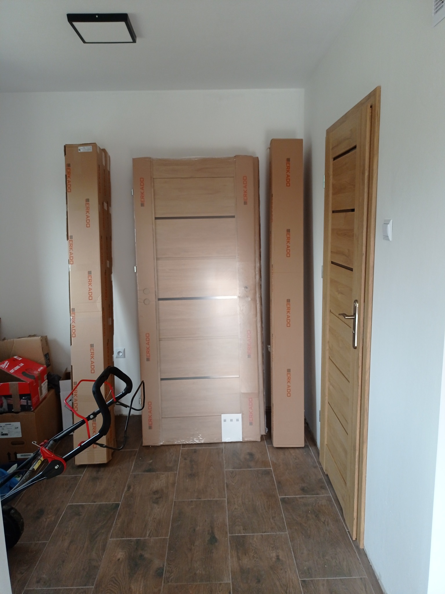 Przygotowane do montażu drewniane drzwi wewnętrzne w opakowaniach, obok zamontowane drzwi z klamką, widoczny wózek transportowy i kartony w tle.