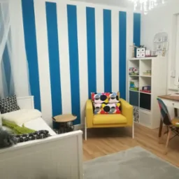 Pokój dziecięcy z biało-niebieskimi pionowymi pasami na ścianie za białym łóżkiem i żółtym fotelem z kolorowymi poduszkami.