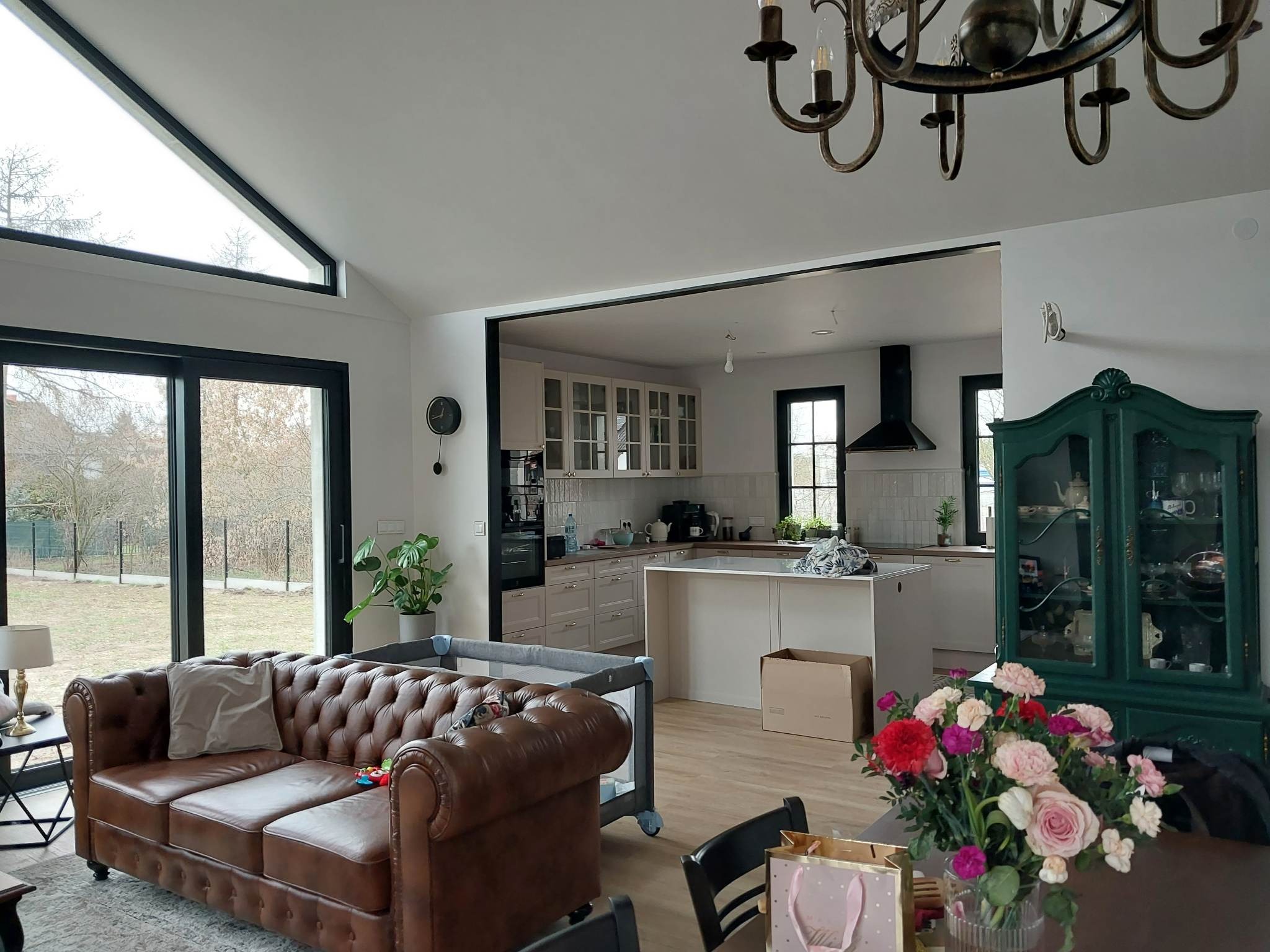 Jasne wnętrze domu po remoncie z widoczną kuchnią z wyspą, brązową skórzaną sofą typu chesterfield i dużym oknem z czarną ramą. W tle widoczny ogród za oknem.