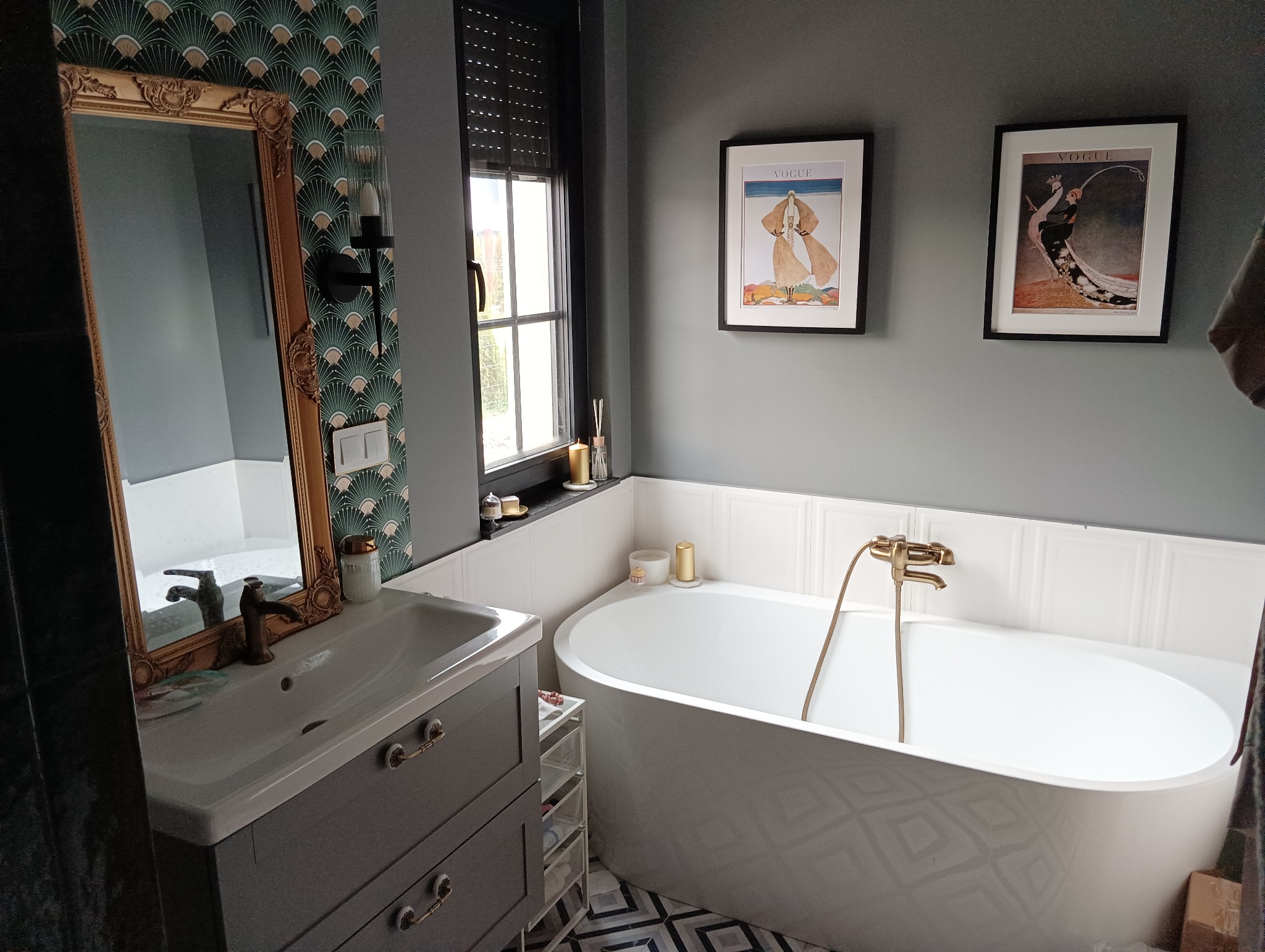 Umywalka z szafką w szarym kolorze, z ozdobnym lustrem w złotej ramie na tle wzorzystej tapety. Obok wanna wolnostojąca z baterią w kolorze złota, okno z żaluzjami i dekoracyjne grafiki na ścianie.