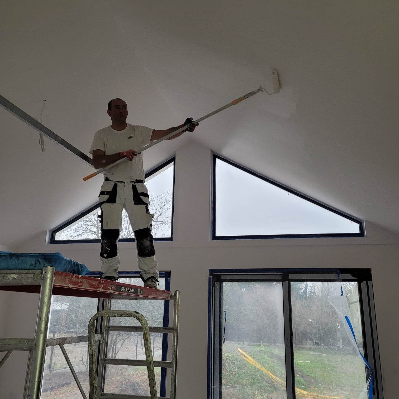 Mężczyzna w roboczym stroju maluje białą farbą sufit na poddaszu za pomocą wałka na teleskopowym kiju, stojąc na rusztowaniu na tle okien o nietypowym, trójkątnym kształcie.