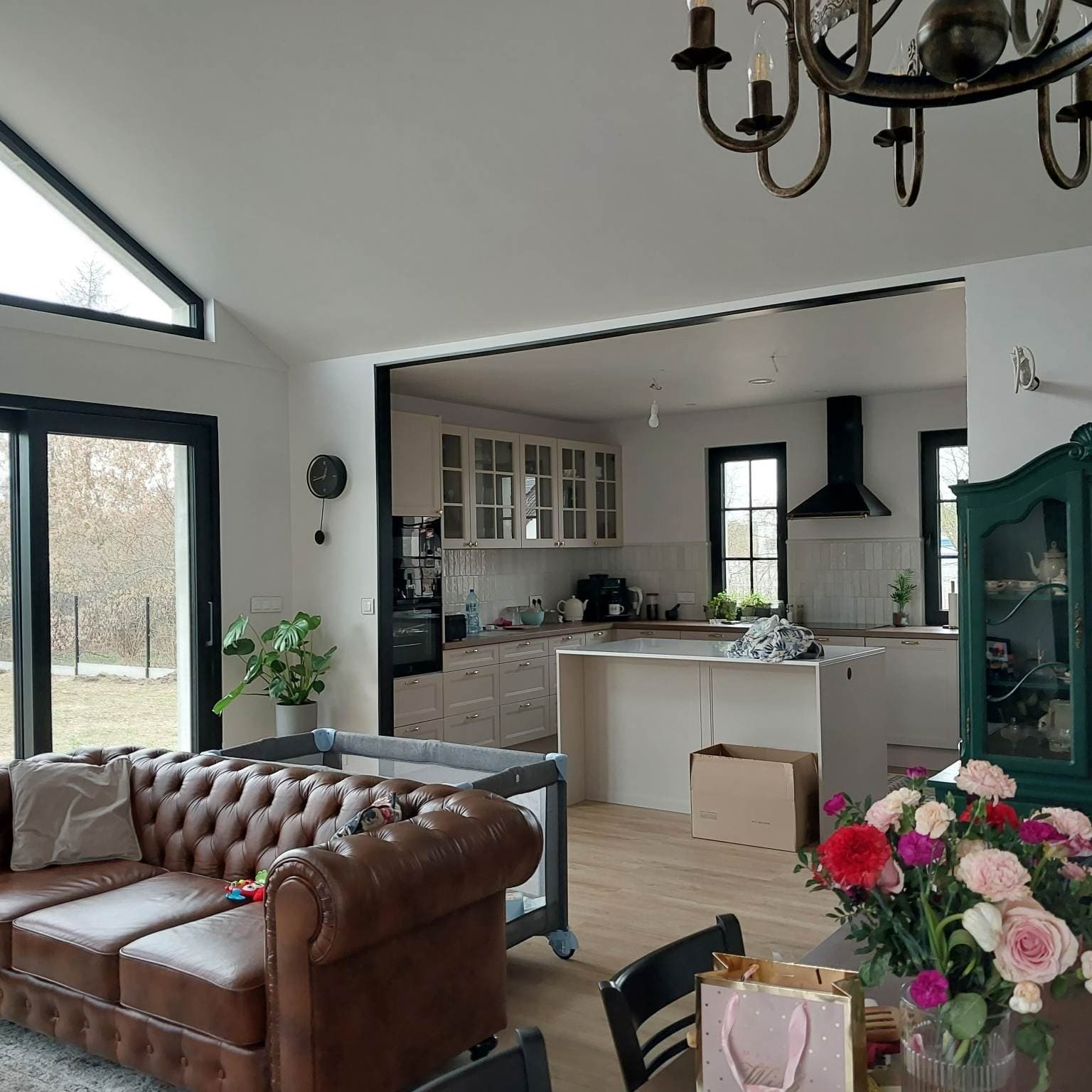 Przestronny salon z otwartą kuchnią w domu jednorodzinnym, widoczny skórzany chesterfield, kwiaty na stole i dziecięce łóżeczko turystyczne, zarys czarnego okapu kuchennego i wyspy kuchennej w tle.