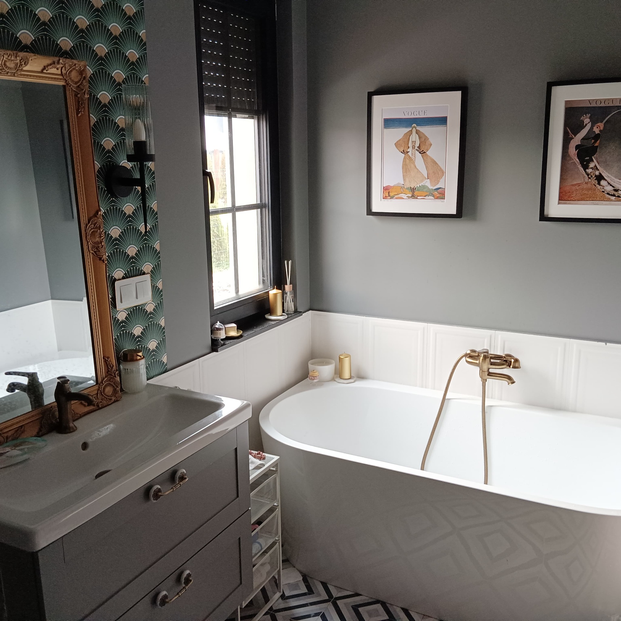 Nowoczesna łazienka w odcieniach szarości z wolnostojącą wanną, armaturą w kolorze starego złota, wzorzystą tapetą i podłogą w geometryczne wzory. Na ścianie wiszą oprawione grafiki.