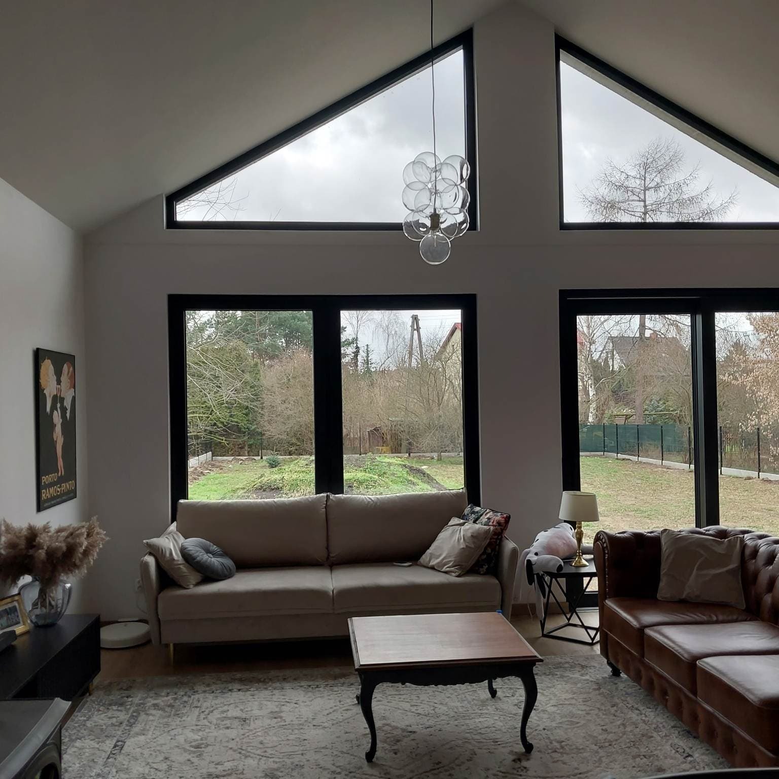 Jasny salon z wysokim sufitem i dużymi oknami w czarnych ramach, widok na ogród, beżowa sofa i brązowa skórzana kanapa, szklany żyrandol.