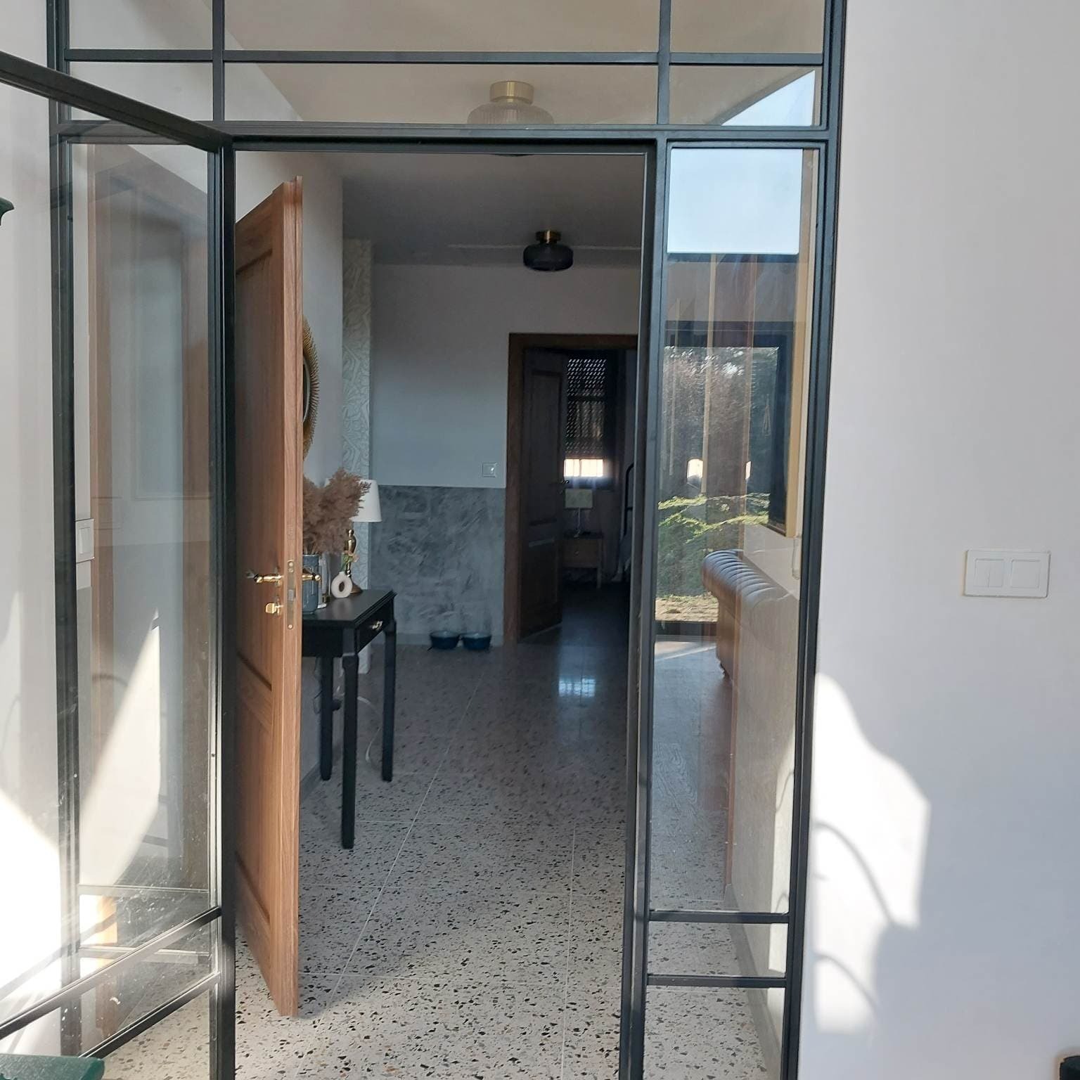 Nowoczesne, przeszklone drzwi wewnętrzne z czarną metalową ramą, widok na przedpokój z lastryko, częściowo otwarte drewniane drzwi po lewej stronie.