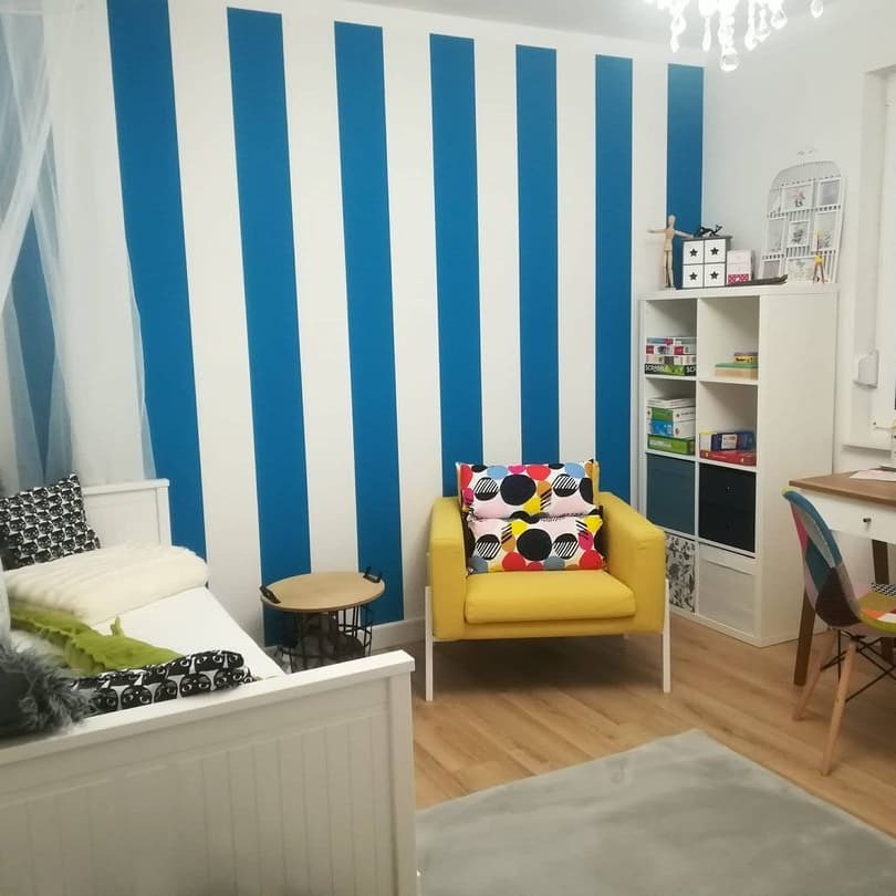 Pokój dziecięcy z biało-niebieskimi pionowymi pasami na ścianie za białym łóżkiem i żółtym fotelem z kolorowymi poduszkami.