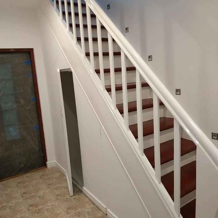 Odnowione schody z białymi balustradami i mahoniowymi stopniami, ściany pomalowane na biało, widoczne gniazdka elektryczne, drzwi zabezpieczone folią malarską.