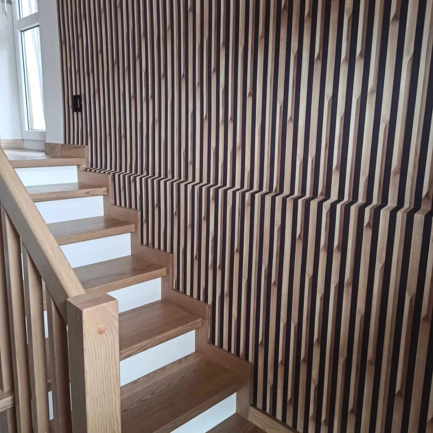 Drewniane schody z balustradą z jasnego drewna, przylegające do ściany z dekoracyjnymi panelami pionowymi w kolorze jasnego drewna i ciemnego brązu, tworzącymi trójwymiarowy efekt.