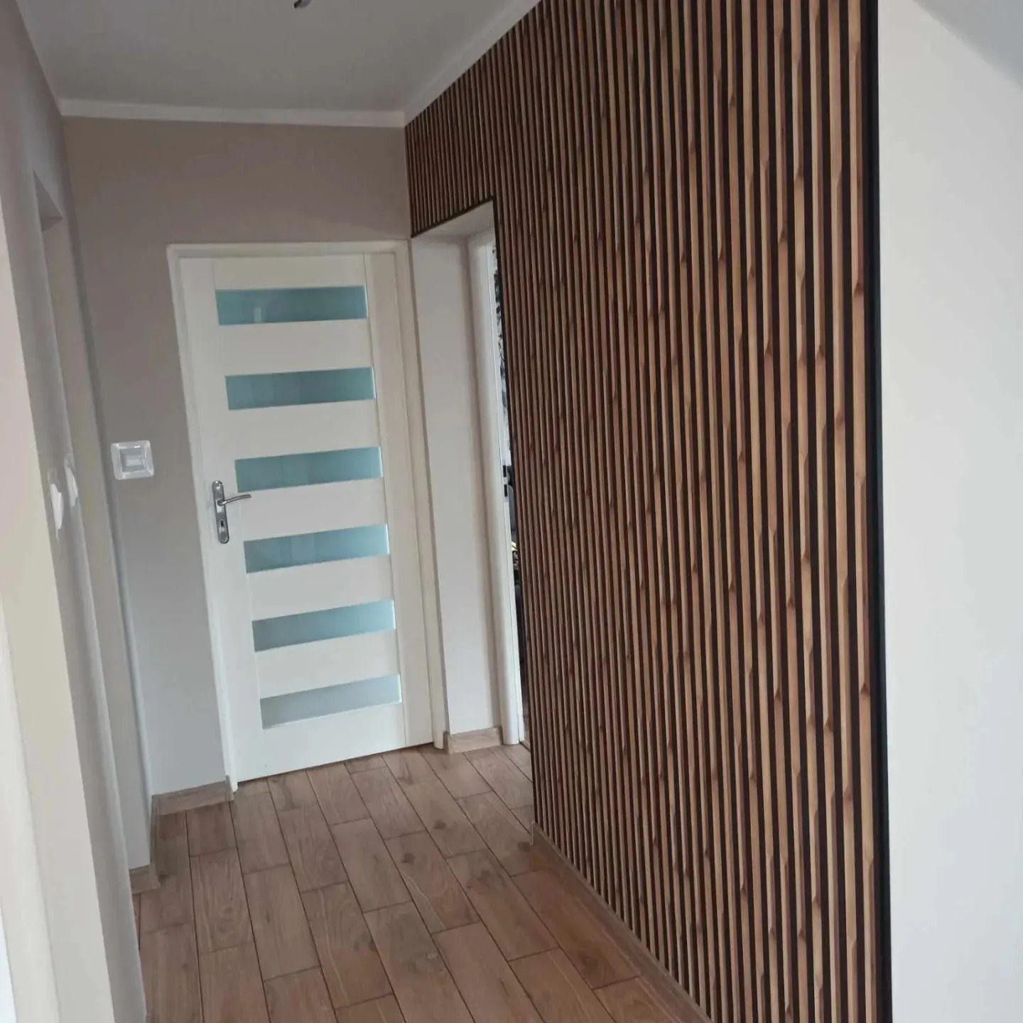 Korytarz po remoncie z drewnianymi panelami lamelowymi na ścianie, jasnymi panelami podłogowymi i białą, przeszkloną wewnętrzną stolarką drzwiową.