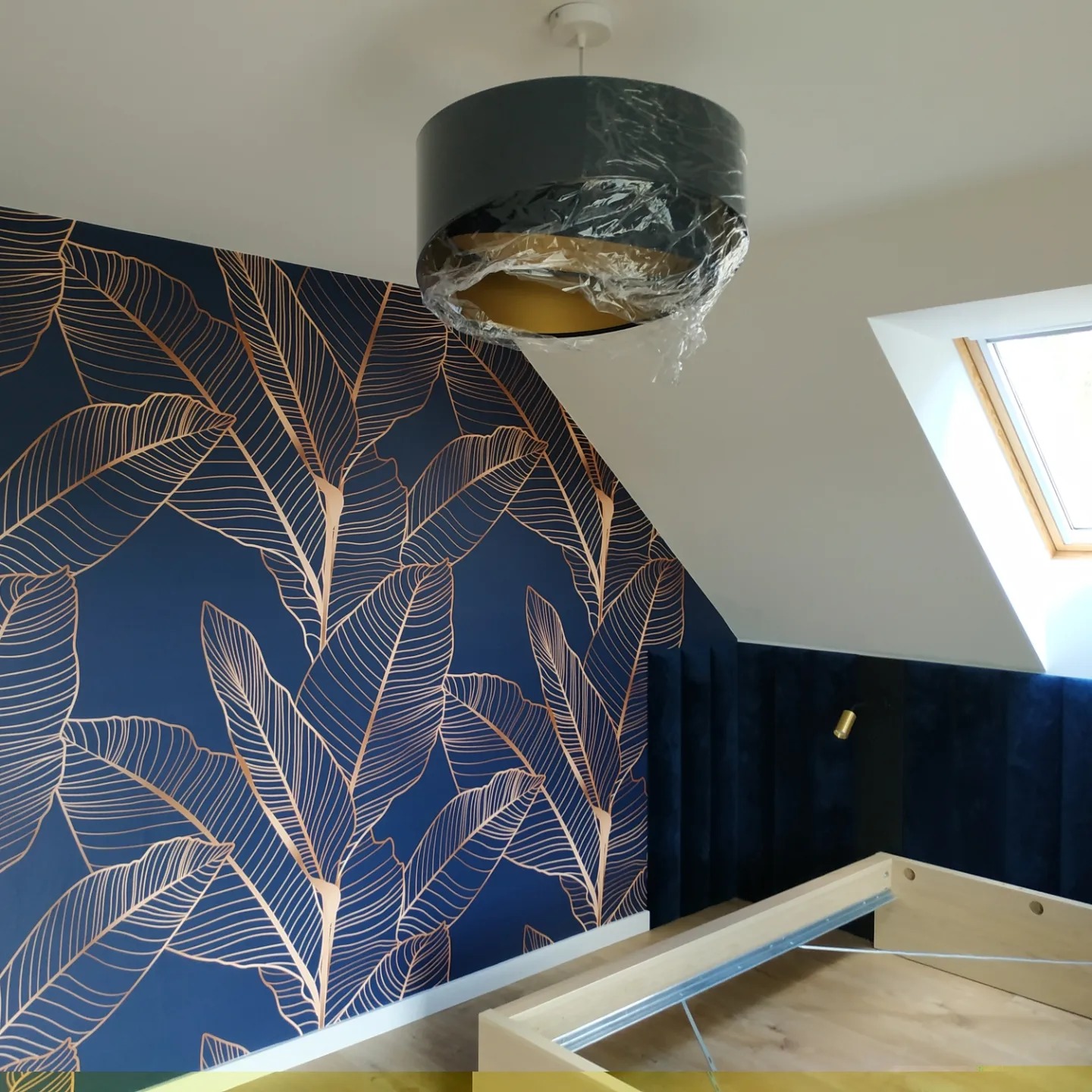 Niebieska ściana z motywem złotych liści, lampa wisząca w folii ochronnej, fragment drewnianej ramy łóżka i okno dachowe w skosie.