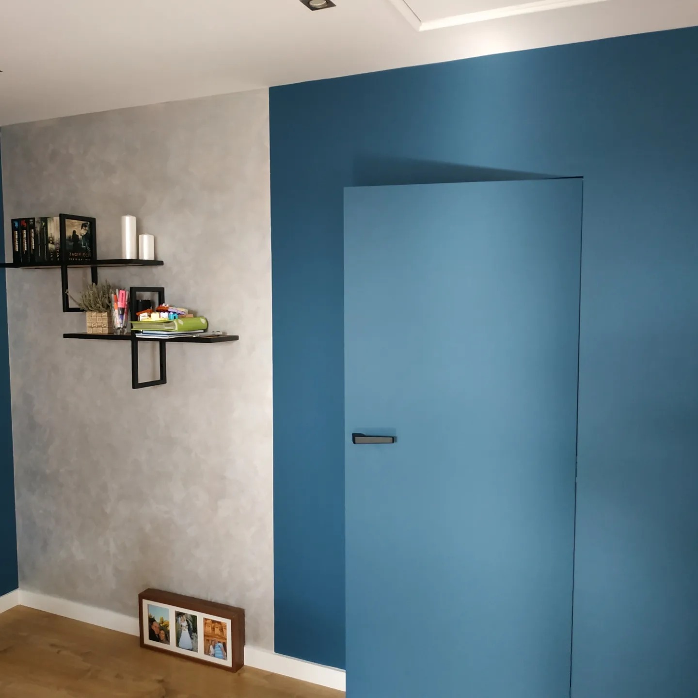 Pokój po remoncie: ściana w odcieniach szarości z dekoracyjną fakturą, kontrastująca z niebieską ścianą z ukrytymi drzwiami. Na ścianie półki z książkami i dekoracjami, na podłodze ramka ze zdjęciami.