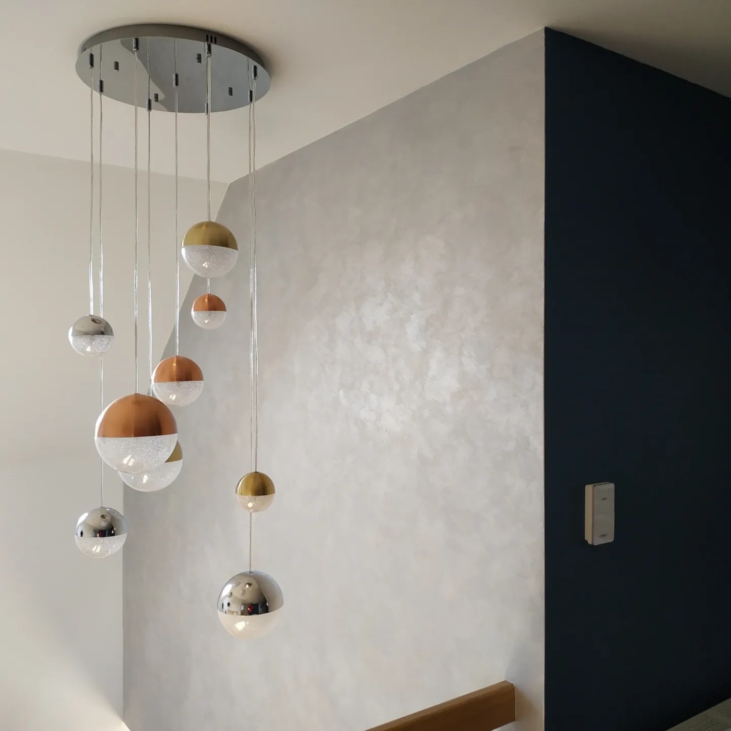 Wielopunktowa lampa wisząca z kulistymi kloszami w kolorach złota, miedzi i srebra, na tle ściany z dekoracyjną, perłową gładzią i fragmentem ściany pomalowanej na ciemny granat.