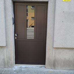 Brązowe drzwi wejściowe z szybą, wkomponowane w szarą elewację budynku. Widoczny fragment ulicy i tabliczka z numerem 27.