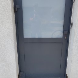 Szare drzwi zewnętrzne z szybą, widoczne zawiasy i zamek. W szybie odbija się osoba robiąca zdjęcie. Na dole kostka brukowa i kratka ściekowa.