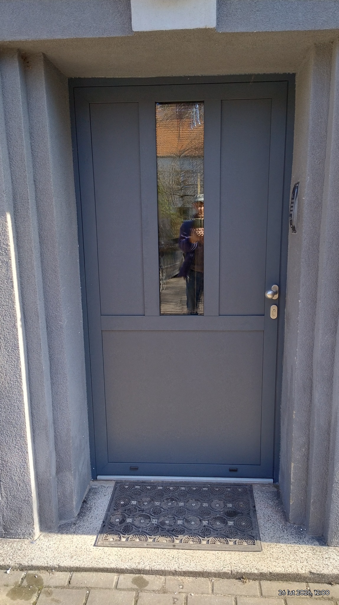 Szare drzwi zewnętrzne z szybą, zamontowane w szarym, teksturowanym murze. Wyraźne odbicie osoby w szybie. Wycieraczka przed drzwiami.