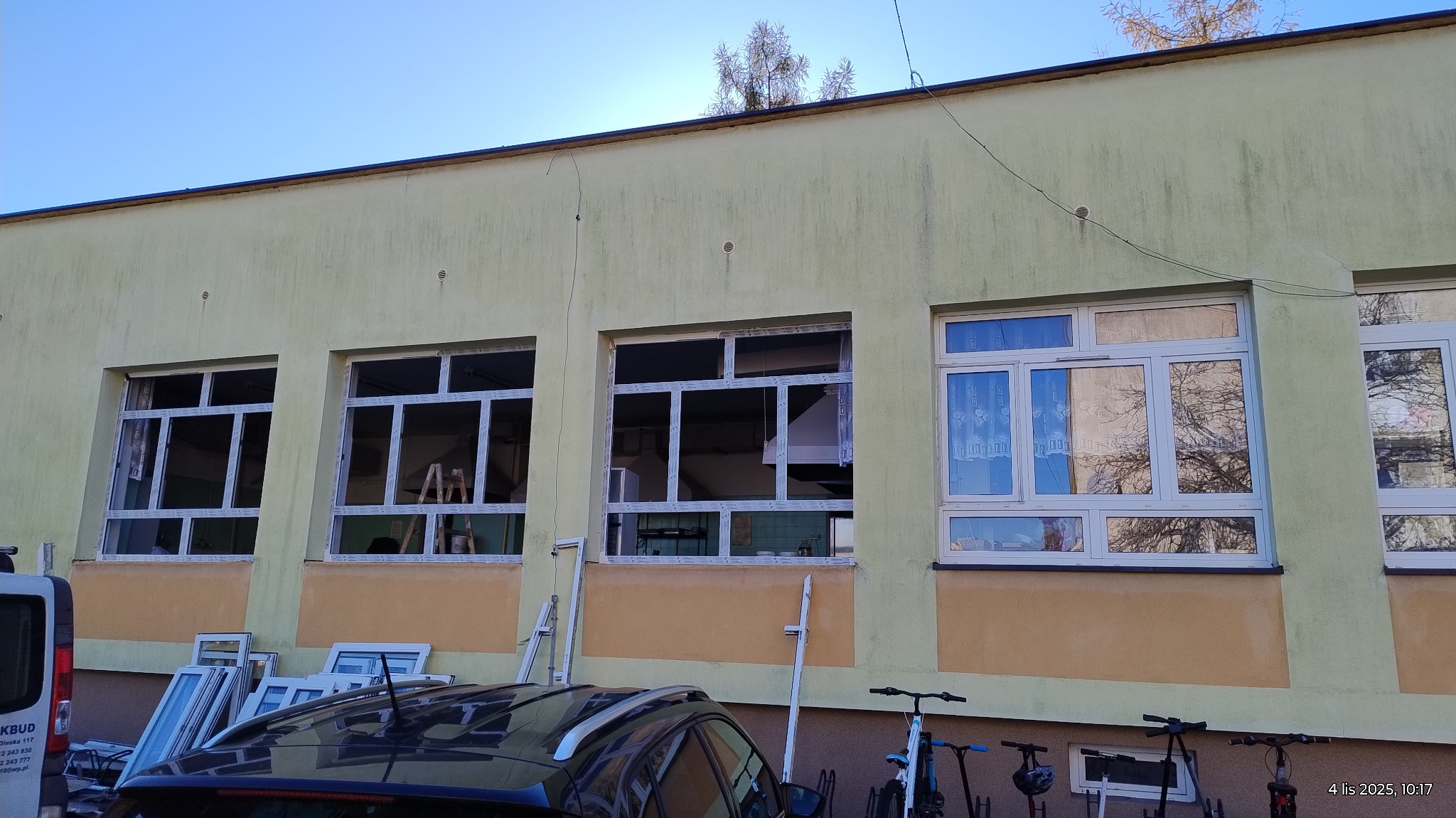 Montaż okien PCV w budynku: widoczne ramy okienne bez szyb, drabina oparta o ścianę, okna przygotowane do montażu obok samochodu. Częściowo zamontowane okno z firanką.