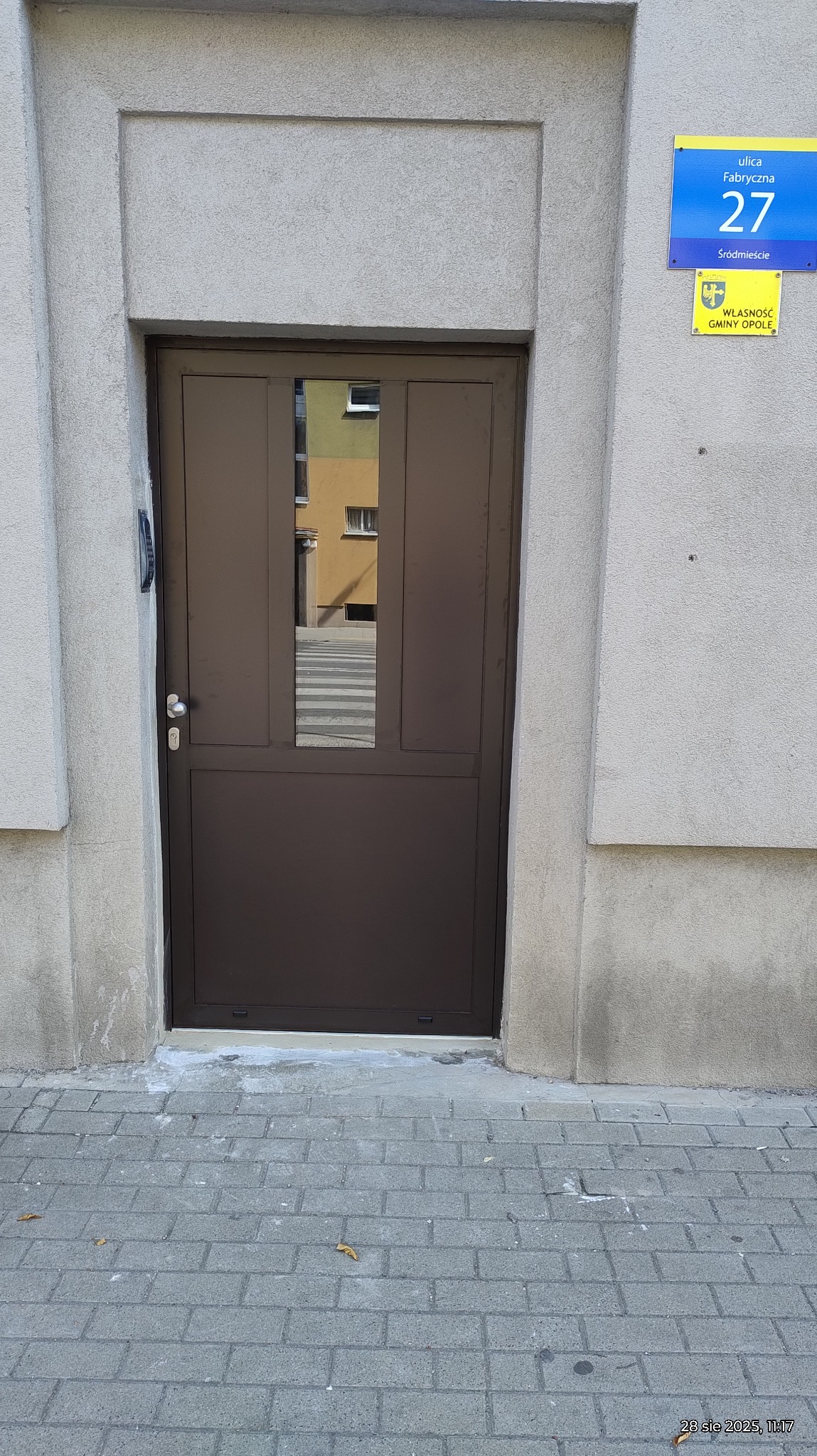 Brązowe drzwi wejściowe z szybą, wkomponowane w szarą elewację budynku. Widoczny fragment ulicy i tabliczka z numerem 27.