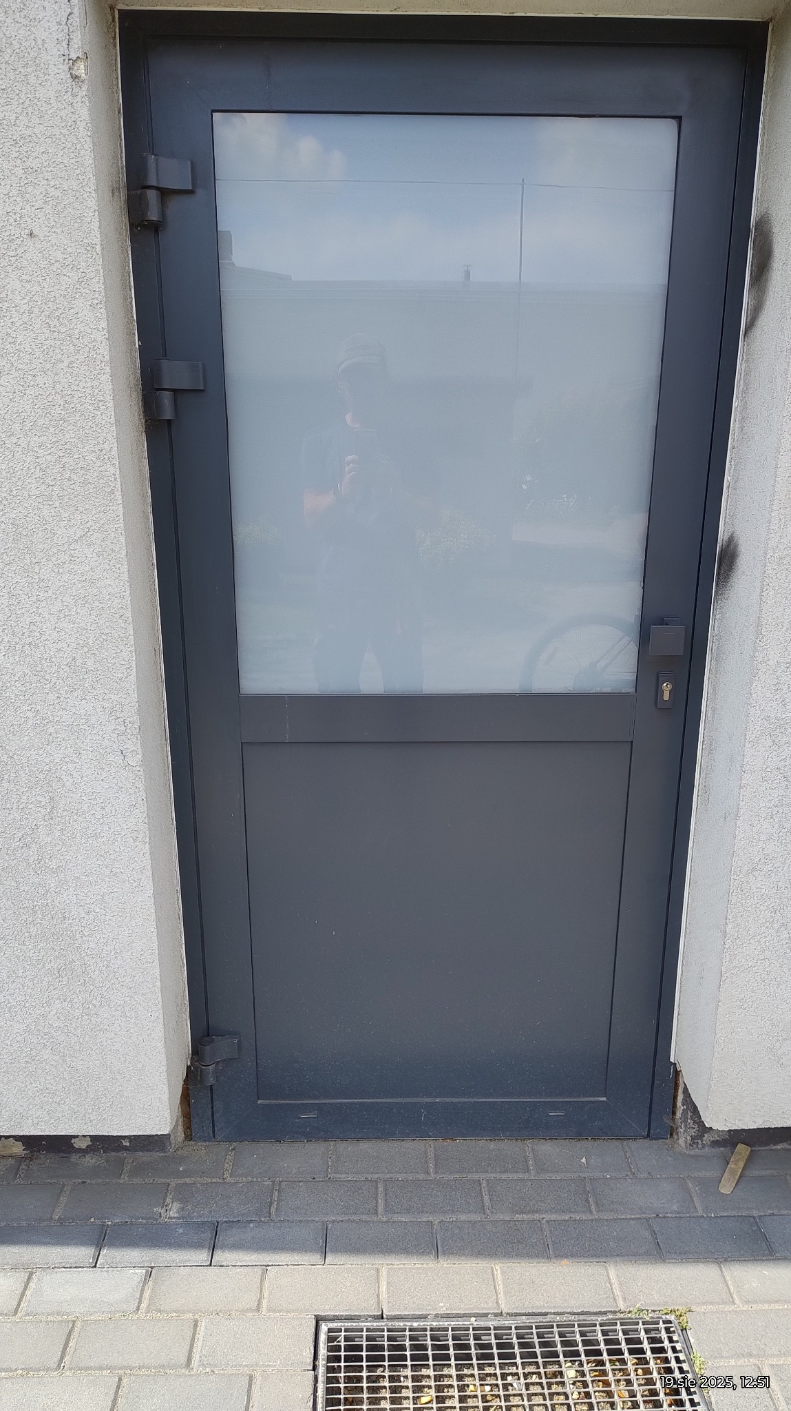 Szare drzwi zewnętrzne z szybą, widoczne zawiasy i zamek. W szybie odbija się osoba robiąca zdjęcie. Na dole kostka brukowa i kratka ściekowa.
