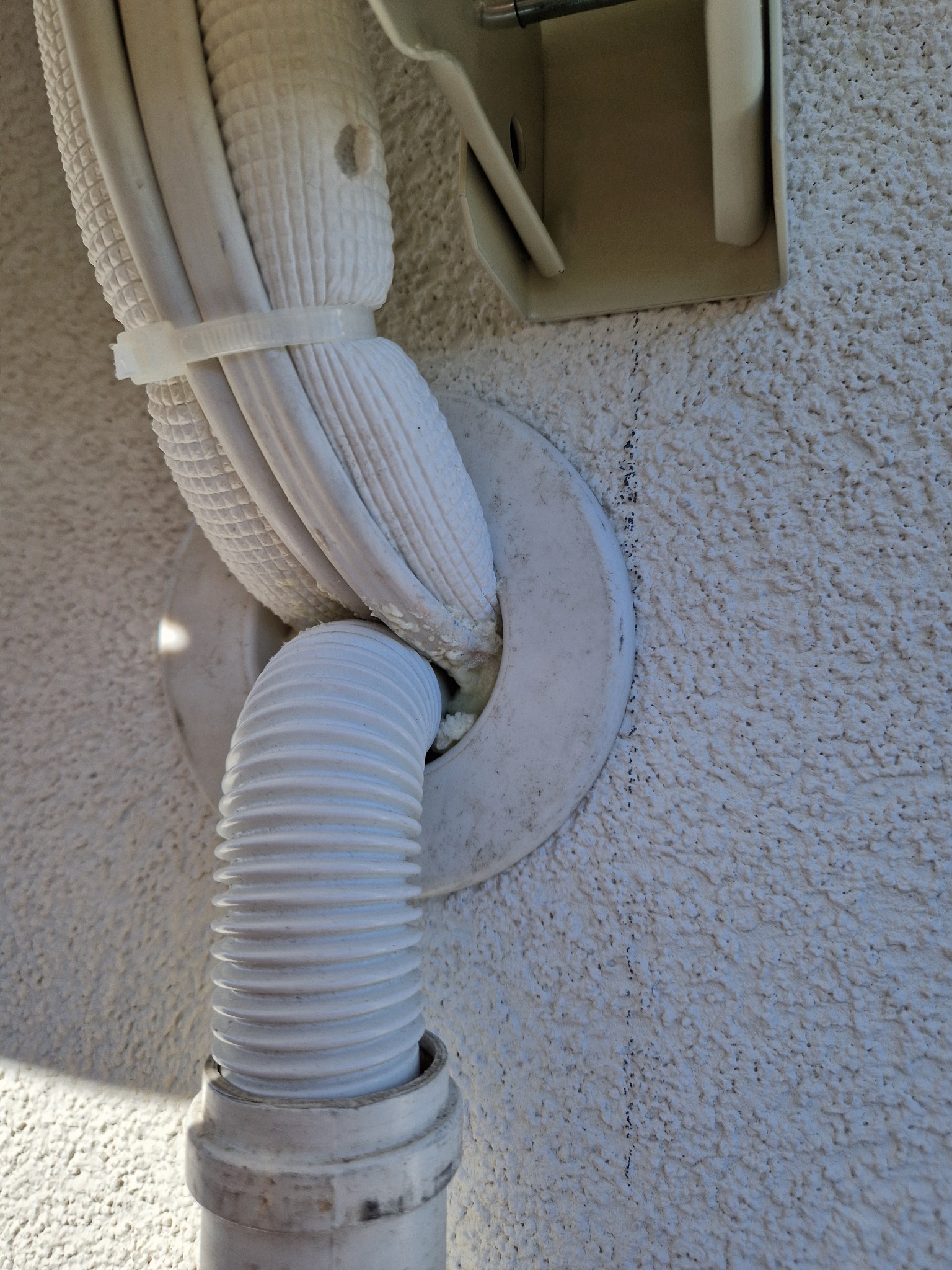 Białe przewody klimatyzacji zabezpieczone opaską zaciskową, wychodzące przez okrągłą uszczelkę w ścianie o szorstkiej fakturze, z widoczną rurą odprowadzającą skropliny.