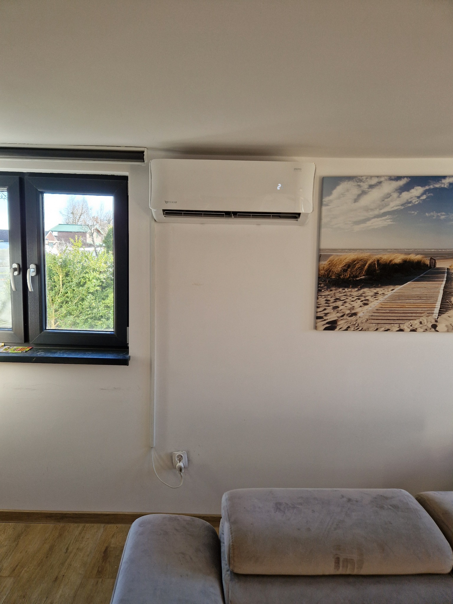 Biały klimatyzator Rotenso zamontowany na ścianie w jasnym pokoju, obok okna z widokiem na zieleń i obrazu z motywem plaży.