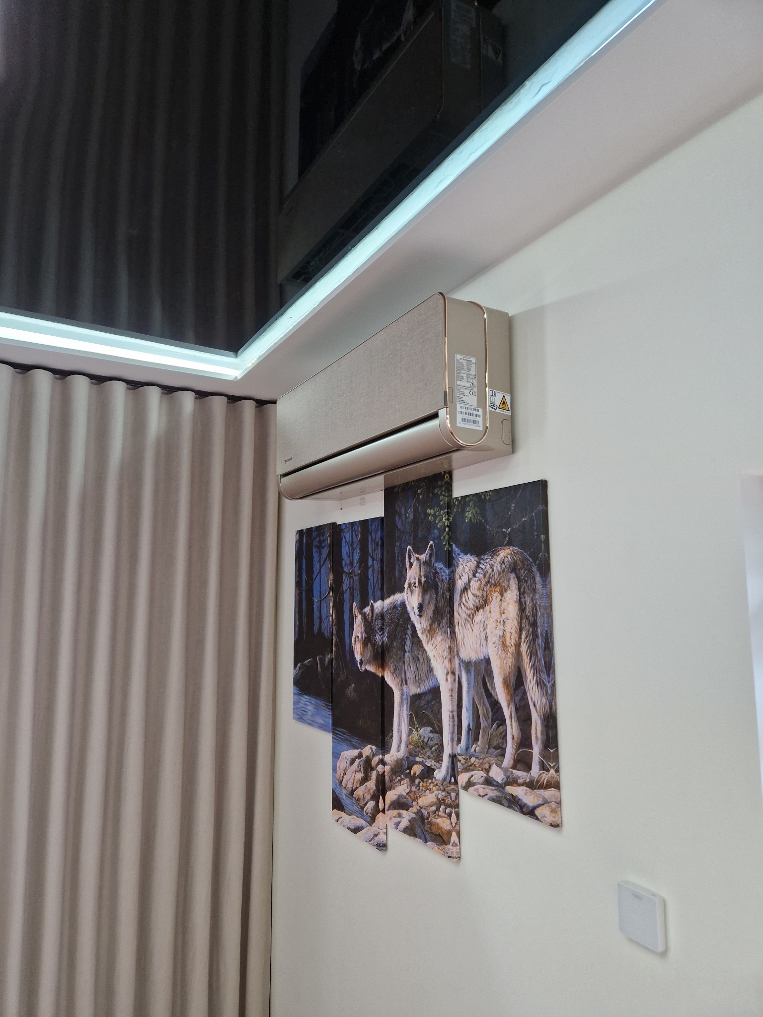 Beżowy klimatyzator ścienny zamontowany w pomieszczeniu z zasłonami i obrazem wilków na ścianie.